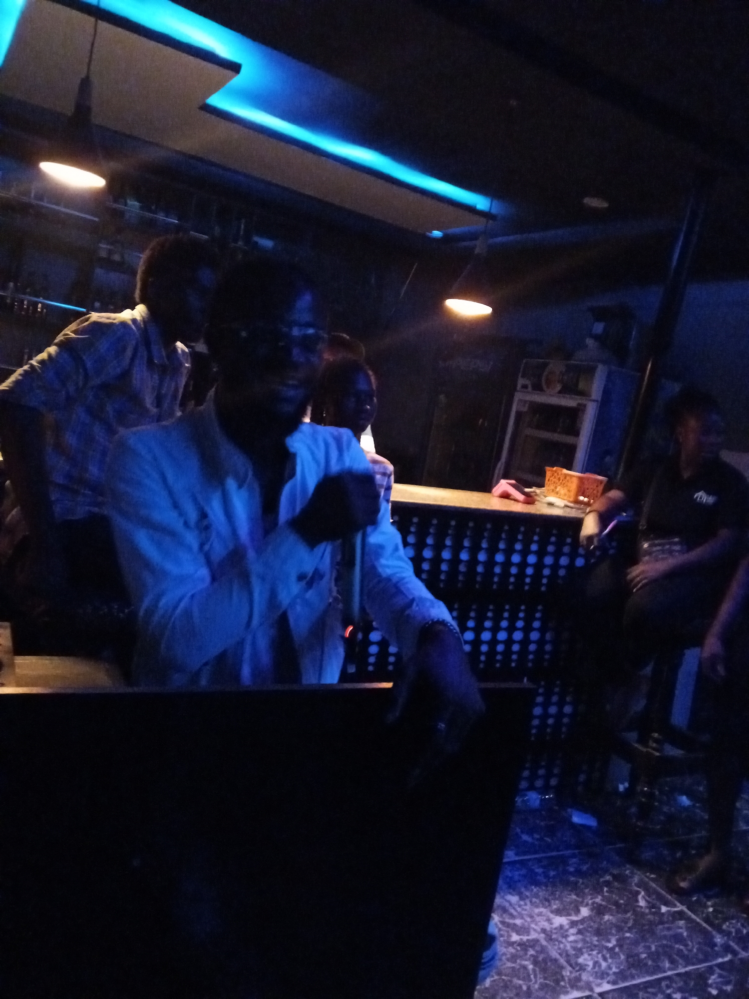 The photo shows a dimly lit bar or club in Lokoja, Nigeria. A man in a light-colored shirt holds a microphone, potentially speaking or singing. Another man in a plaid shirt stands behind him. A bar with patterned front is visible, and a woman sits on a stool behind the bar. There are lights on the ceiling and under the bar. The floor is tiled with a marble-like pattern.