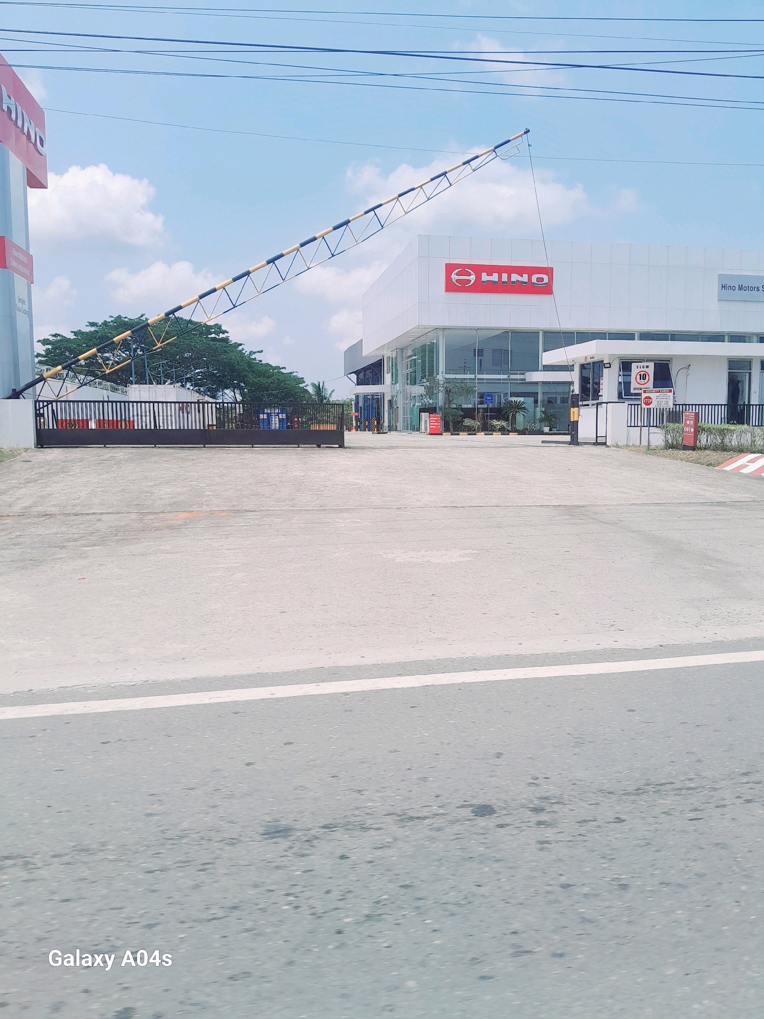 The image shows the entrance to a Hino Motors facility in Balikpapan, Indonesia. The entrance features a large black gate with a yellow and black striped barrier arm extending over the roadway. To the left of the gate is a tall blue and white sign with red lettering that reads "HINO." The Hino Motors building is visible behind the gate, with large glass windows and a red Hino logo. A small guardhouse is located to the right of the entrance, with a sign indicating a speed limit of 10 km/h and "SECURITY CHECK." The ground in front of the gate is paved with concrete. The sky is blue with scattered white clouds, and there are several power lines crossing the top of the frame. The overall impression is of a commercial vehicle dealership or service center.
