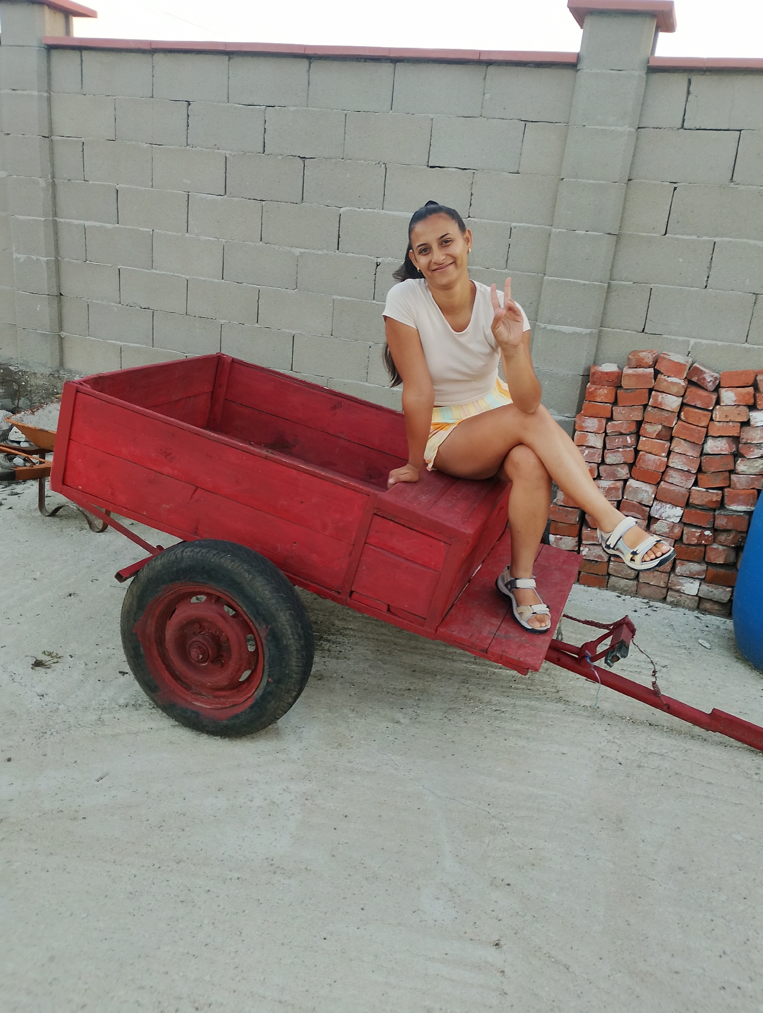 A woman is sitting on a red wooden cart with a single wheel in front, in Zimnitsa, Bulgaria. The cart is positioned on a light grey concrete surface. Behind the cart is a wall constructed from light grey cinder blocks. To the right of the cart, a stack of red bricks is visible, and a large blue cylindrical object is partially in frame. The woman is wearing a light-colored t-shirt and a patterned skirt, and she has her legs crossed, with her left hand resting on the cart and her right hand raised in a peace sign. The background shows the upper section of the cinder block wall with a red-tiled coping. The overall scene appears to be an outdoor area, possibly a yard or construction site.