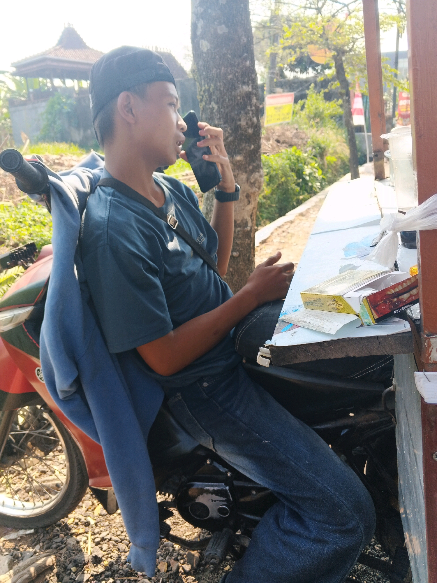 A young man, appearing to be of Indonesian descent, is seated on a motorcycle and talking on a smartphone. He is wearing a dark blue t-shirt, blue jeans, and a black baseball cap. A light blue jacket is draped over the back of the motorcycle. In the foreground, a red motorcycle is visible. The man is sitting at a wooden table where a pack of cigarettes is displayed. The background shows a building with a traditional Indonesian roof, trees, and foliage, suggesting a semi-rural or suburban setting within Kalirejo, Indonesia. The lighting indicates it is daytime.
