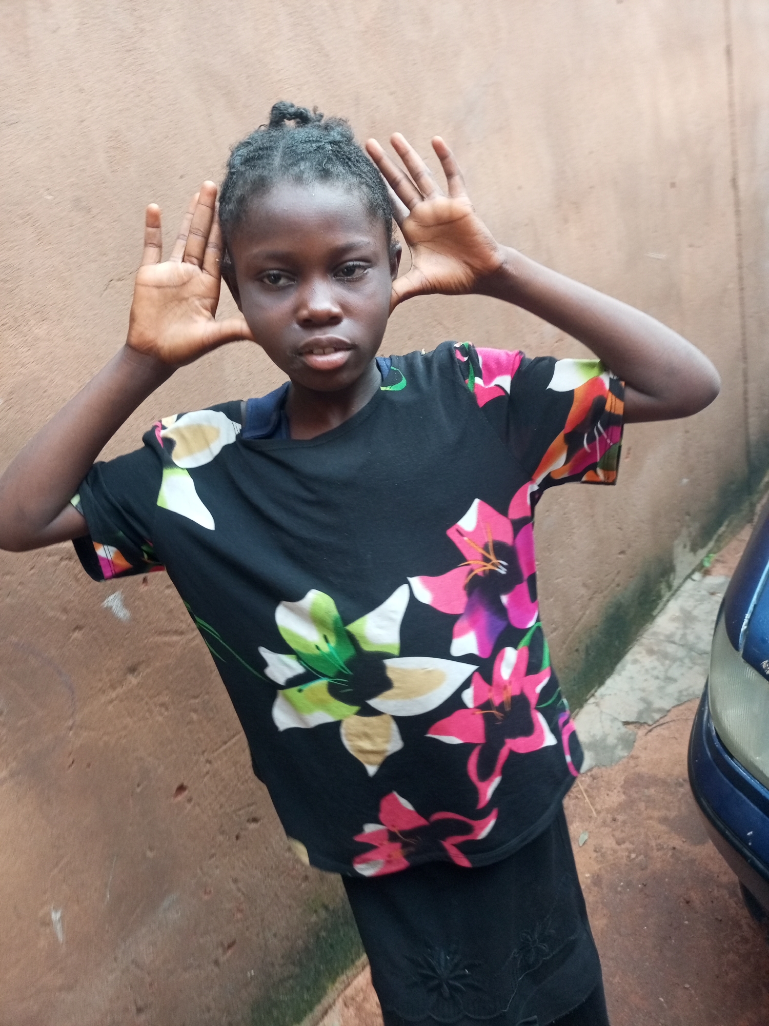 A young Black female stands outdoors in front of a textured brown wall. She is looking directly at the camera with an open mouth and her hands raised to her temples, palms facing forward, with fingers splayed. She has dark, braided hair pulled up in a bun. She is wearing a short-sleeved black t-shirt with a colorful floral print featuring large pink, white, and yellow flowers with green accents. Below the t-shirt, she is wearing a black skirt. To the right of the frame, the front quarter panel and headlight of a dark blue vehicle are visible. The ground appears to be concrete or paved with some cracks. The overall lighting suggests it is daytime. The location is an outdoor setting, possibly in the city of Tempure, Nigeria, based on the provided context.