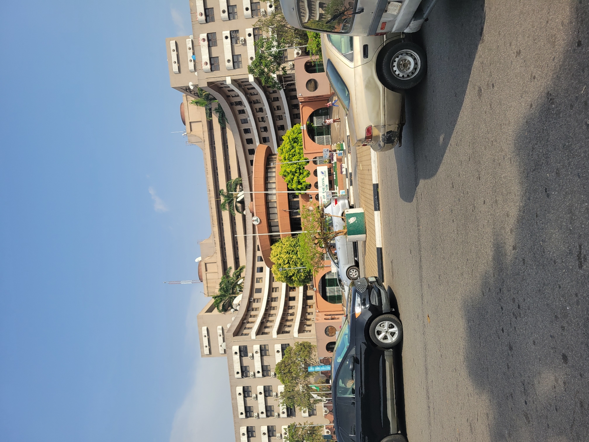 The image captures a street scene in Abuja, Nigeria. A curved, multi-story building with a light brown exterior is visible, featuring rows of windows and balconies. A shorter, reddish-brown structure is in front of the taller building, with arched doorways and green foliage. Several cars are parked along the side of the road, including a black SUV and a white van. Sidewalks are visible along the road. The sky is mostly clear and blue. People are walking near the entrance of the building.