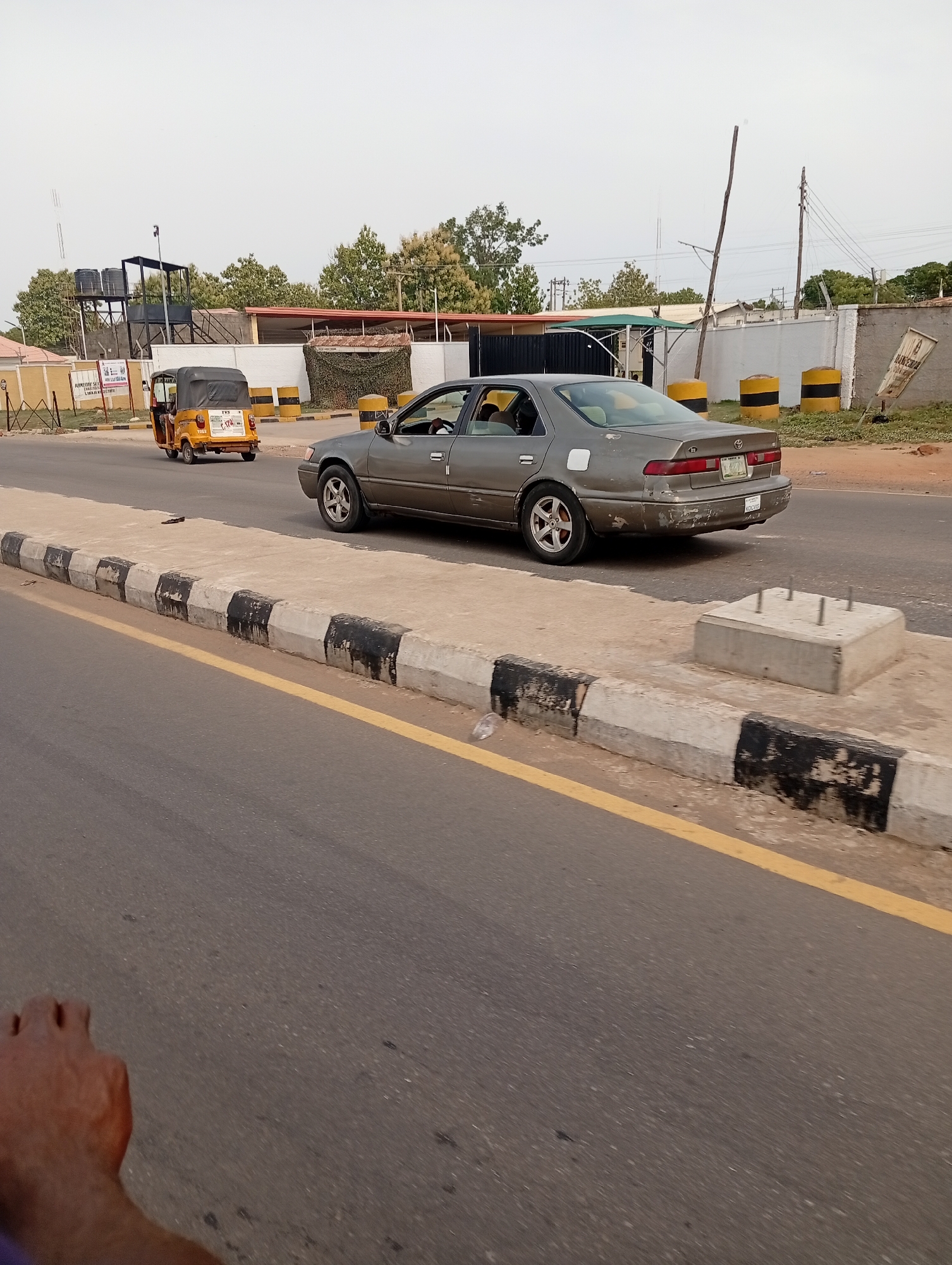 The image, taken in Adankolo, Nigeria, shows a gray Toyota Camry driving on a road next to a curb painted with alternating black and white stripes and a yellow line. A yellow auto rickshaw is traveling in the same direction in the other lane. In the background is a building with camouflage netting. Along the side of the road are concrete blocks painted with black and yellow stripes. A concrete base with metal posts sticking out of it sits along the curb. A portion of a person's arm is visible in the lower left corner of the image.