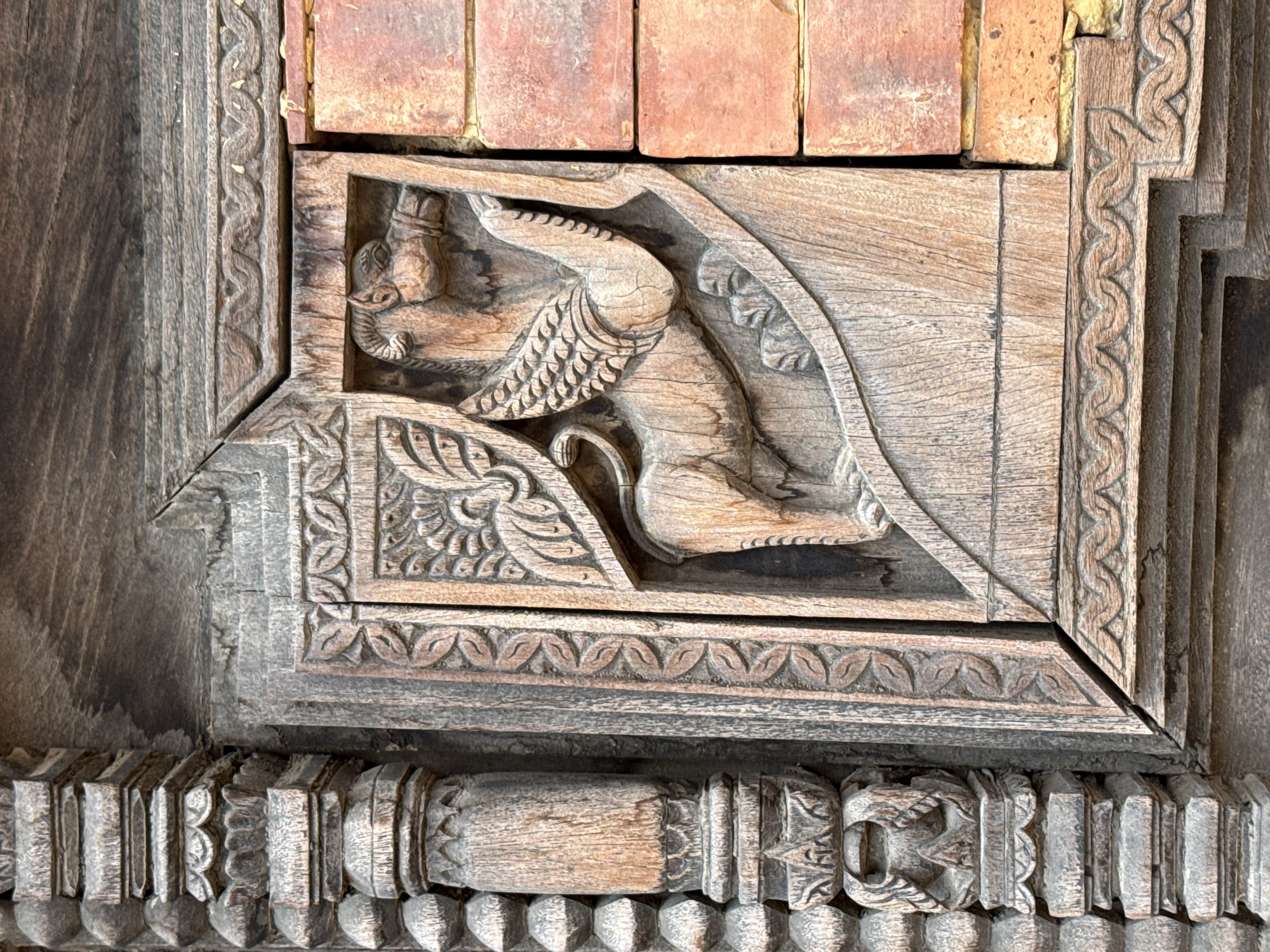 The image shows a detailed wood carving, possibly from a structure in Bhaktapur, Nepal. The carving features a mythical creature, perhaps a Garuda or a similar winged figure with an elephantine trunk, kneeling or crouching. The creature has wings and a tail, and is depicted in a dynamic pose. The background and surrounding frame of the carving show intricate patterns and floral motifs, indicating traditional craftsmanship. The wood is weathered and appears to be aged. The carving is adjacent to brickwork. The overall setting suggests an architectural detail from a temple, palace, or other significant building in Bhaktapur.