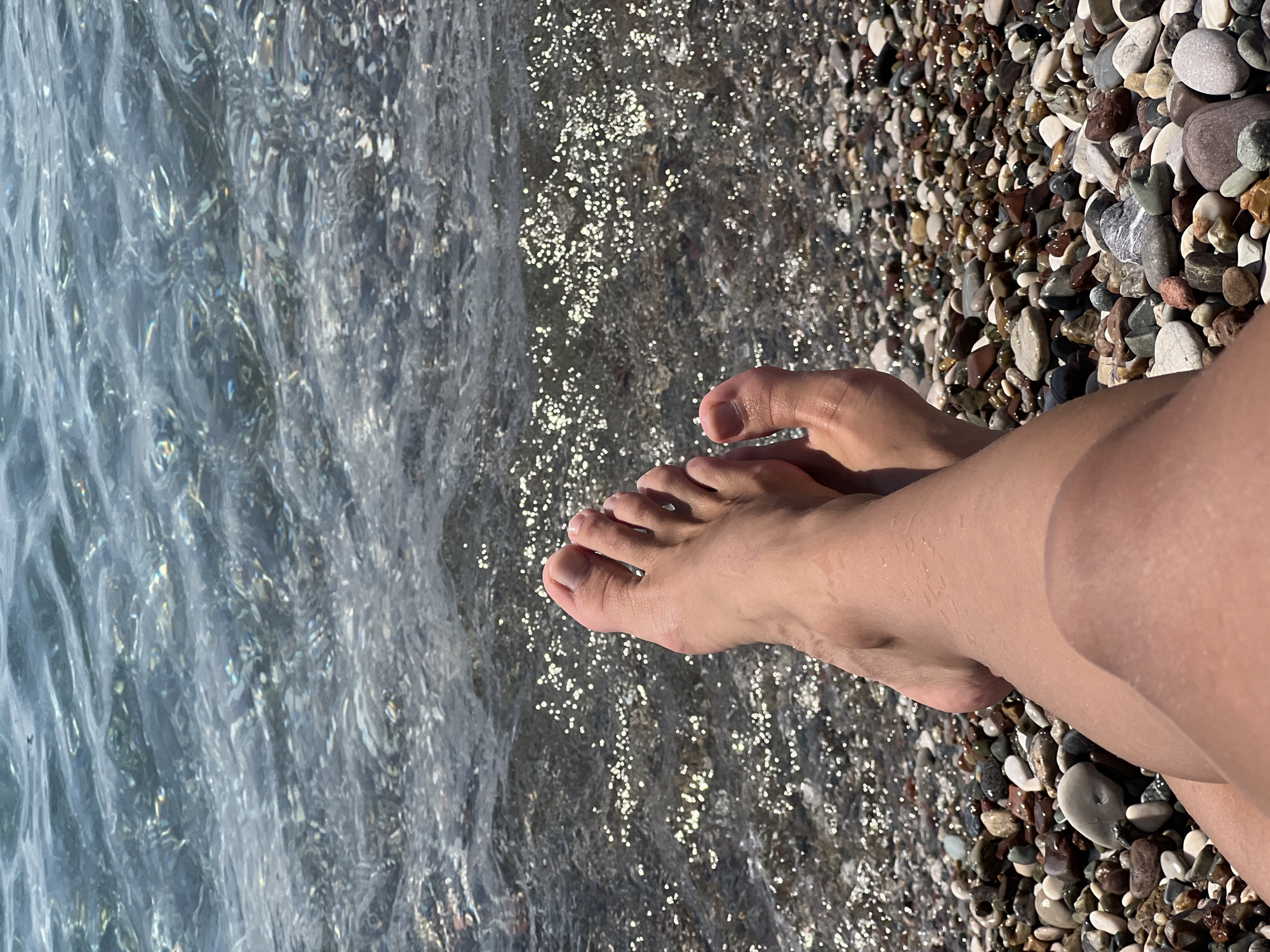 The image captures a close-up, overhead view of a person's bare feet and lower legs resting on a pebble beach. The water of the Mediterranean Sea, with its characteristic clear turquoise hue and shimmering surface from sunlight, laps gently at the shore. The pebbles on the beach are a mix of sizes and colors, predominantly grey, brown, and white, with some exhibiting smooth, rounded shapes. The skin on the legs and feet appears wet, indicating recent immersion or splashes from the sea. The lighting suggests it is daytime, likely under bright sunlight. The specific location within Antalya, Turkey, is not discernible from the image itself, but the scene is consistent with any of Antalya's numerous pebble beaches lining the Mediterranean coast.