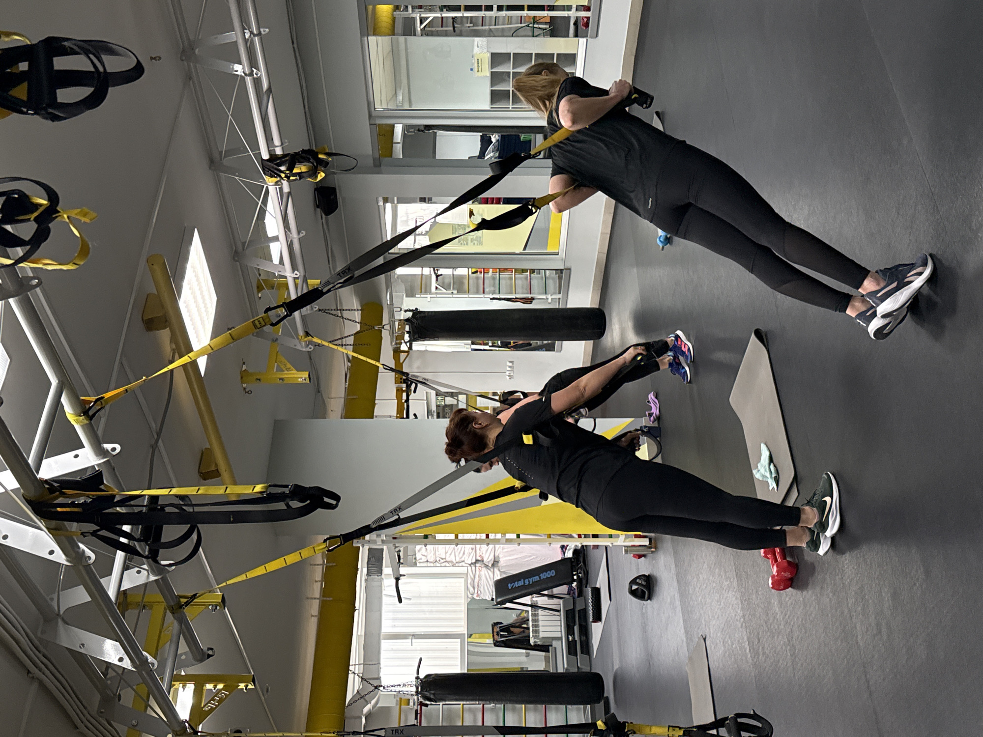 The image shows two women performing a TRX exercise in what appears to be a gym or fitness studio. Both women are dressed in black athletic wear. The woman in the foreground has her hair pulled back in a bun. They are using yellow and black TRX suspension training equipment. The gym has a black floor, white walls, and metal support beams on the ceiling. Gym equipment such as a Total Gym 1000, dumbbells, and a punching bag are visible in the background. The location is likely an indoor fitness facility. There is nothing in the image to indicate that the location is Chaikovsky, Russia.