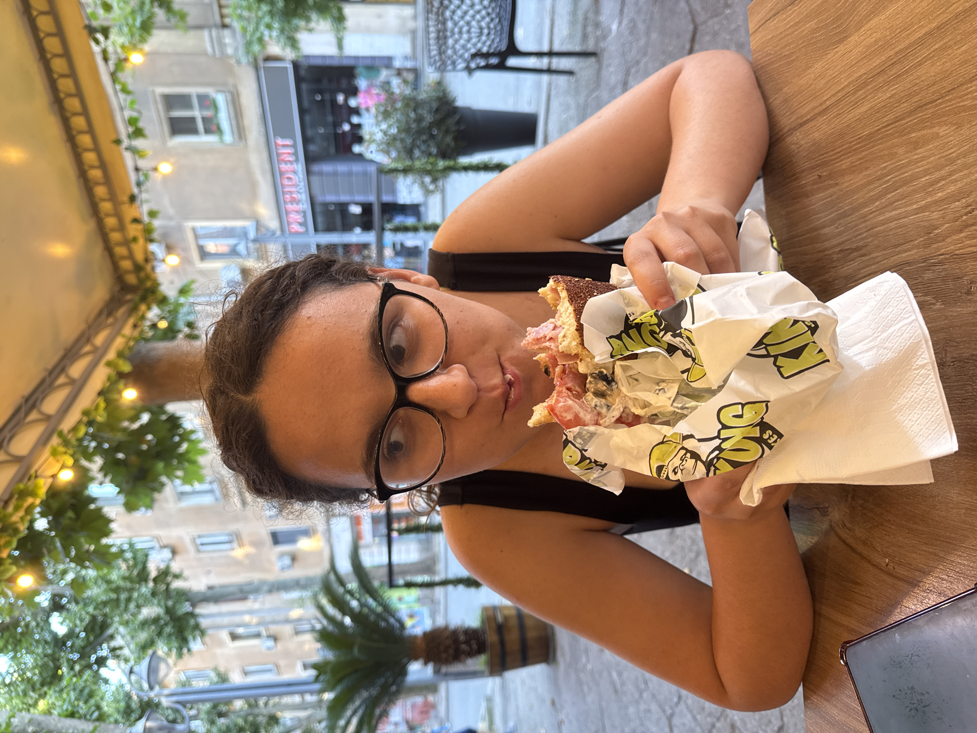 A young woman with dark curly hair and black-rimmed glasses is seated at a wooden table, eating a large sandwich wrapped in paper. She is wearing a black tank top and is looking off to the side, with a piece of the sandwich in her mouth. The sandwich appears to be made with a dark, seeded bun and is filled with meat, vegetables, and sauce. The paper wrapper has a cartoon character and text on it. In the background, there is a building with "PRESIDENT" signage and several potted plants. The scene appears to be outdoors, possibly at a cafe or restaurant in Burgas, Bulgaria, as indicated by the architecture and vegetation. The overall atmosphere is casual and relaxed.