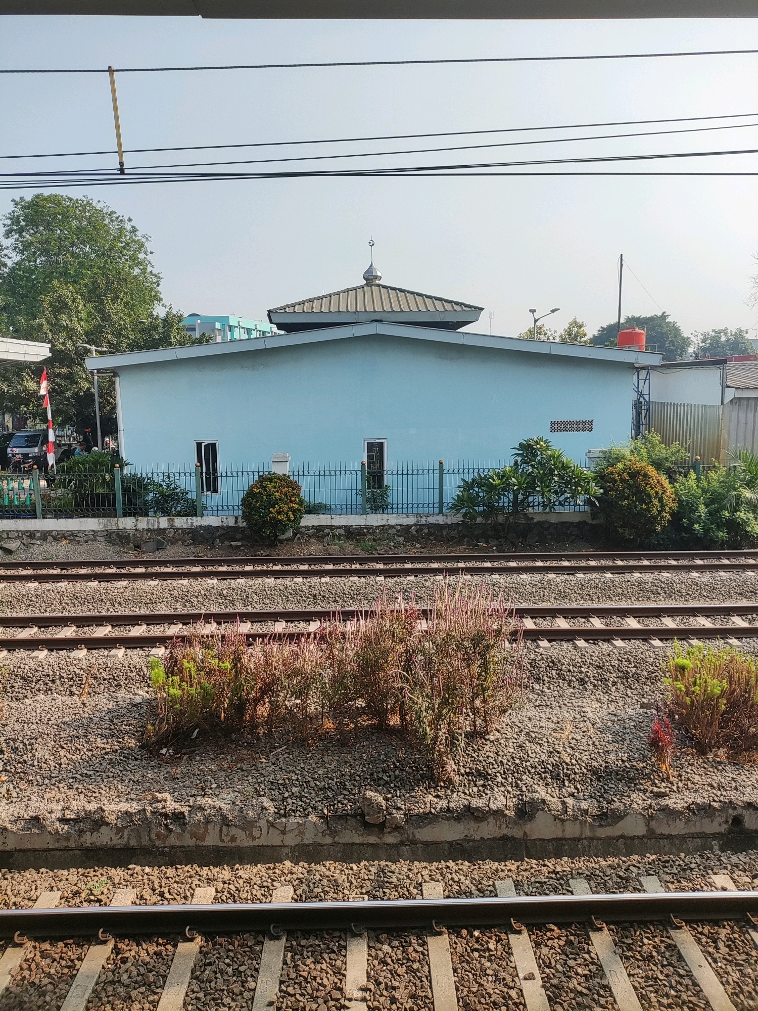 The image shows a light blue building, likely a mosque given the crescent moon on its roof, situated behind a black metal fence. The building has a gabled roof and a few windows. Adjacent to the building, there is greenery and a red water tank on a raised platform. In the foreground, there are multiple sets of train tracks laid on gravel. Some dry, reddish plants are growing between the tracks. The sky is clear and blue, with some power lines running across the top of the frame. The overall scene appears to be an outdoor location, possibly a train station area, in East Jakarta, Indonesia.