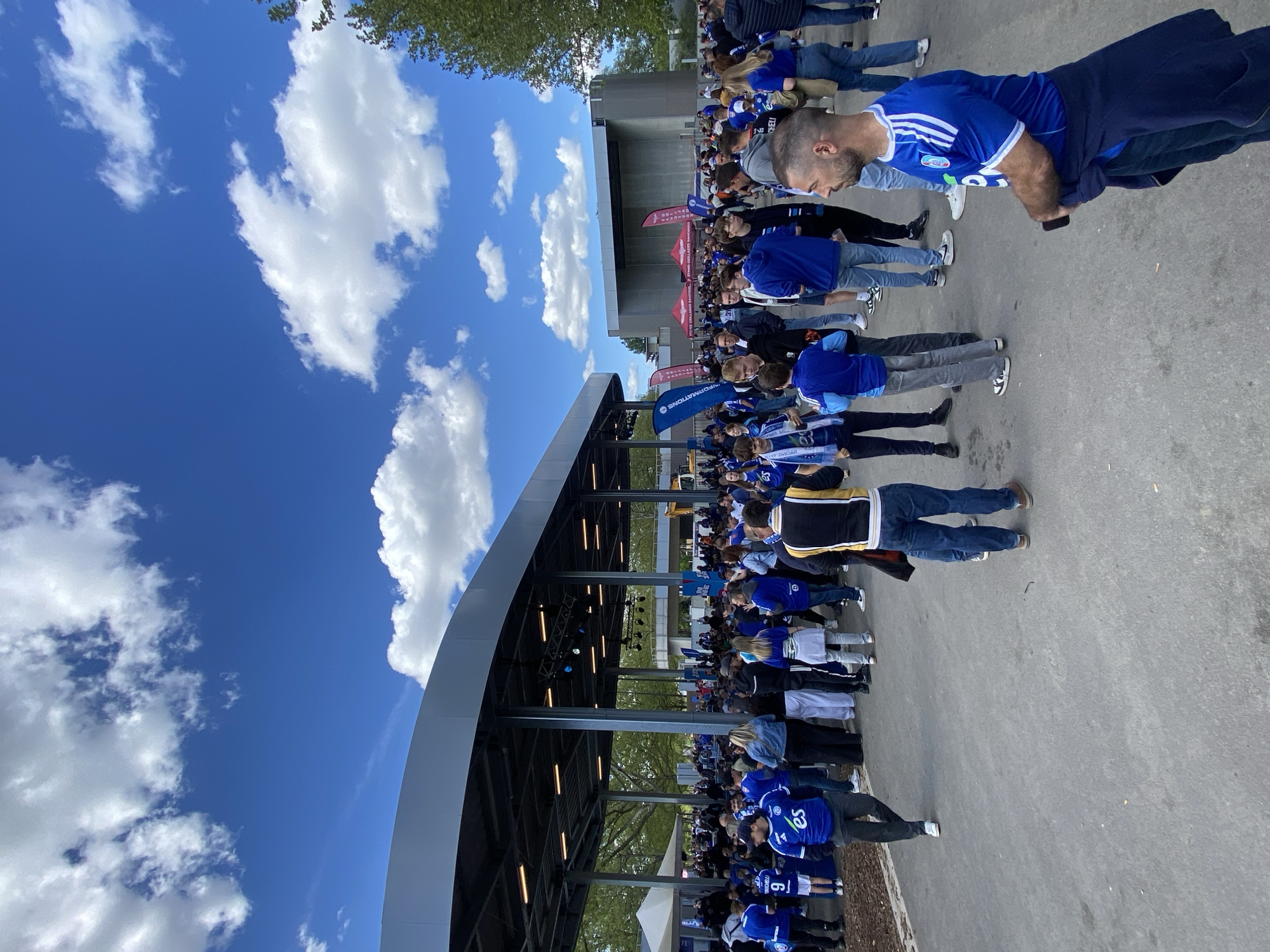 A crowd of people is gathered outside a building under an arched roof. Most of the people are wearing blue clothing, possibly team jerseys. The building is constructed with metal supports and lighting fixtures under the roof. The ground is paved, likely a concrete surface. Overhead, there is a blue sky with scattered white clouds. This scene is likely in Strasbourg, France, based on the team colors worn by many people in the crowd.