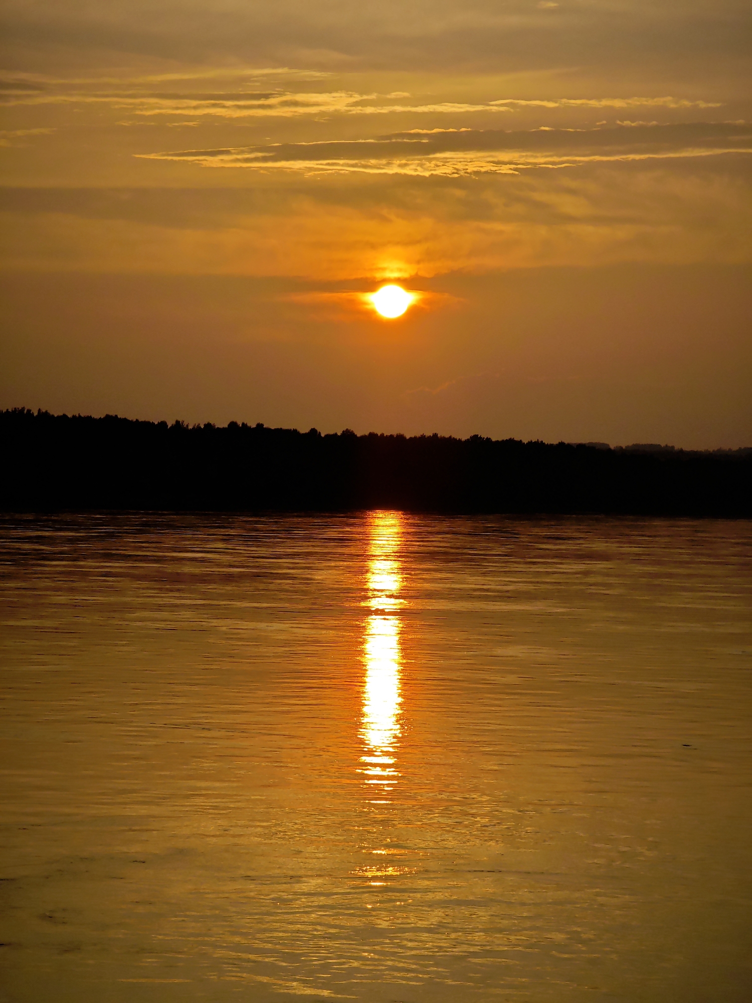 The image depicts a sunset over a body of water, likely a river or lake, near Maloe Nakhval'skoe, Russia. The sky is filled with horizontal clouds illuminated by the setting sun, which appears as a bright, circular orb near the center of the horizon. A distinct reflection of the sun creates a shimmering path across the water's surface. The shoreline is silhouetted against the sky, forming a dark, tree-lined band. The overall scene is bathed in warm, golden and orange hues characteristic of a sunset. No people or specific man-made structures are visible, focusing the image on the natural landscape.
