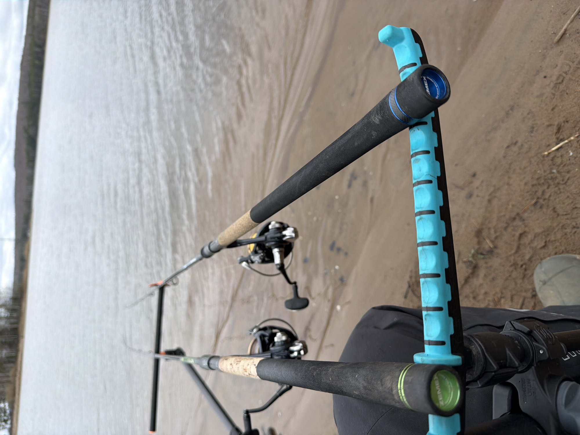 A ground-level, medium-distance shot depicts two fishing rods propped up on a stand on a sandy beach in Chaikovsky, Russia. The rods are angled diagonally across the frame, with the handles closest to the viewer. Fishing reels are attached to each rod. A blue object is visible near the rod handles. The background consists of a body of water and vegetation along the shoreline.