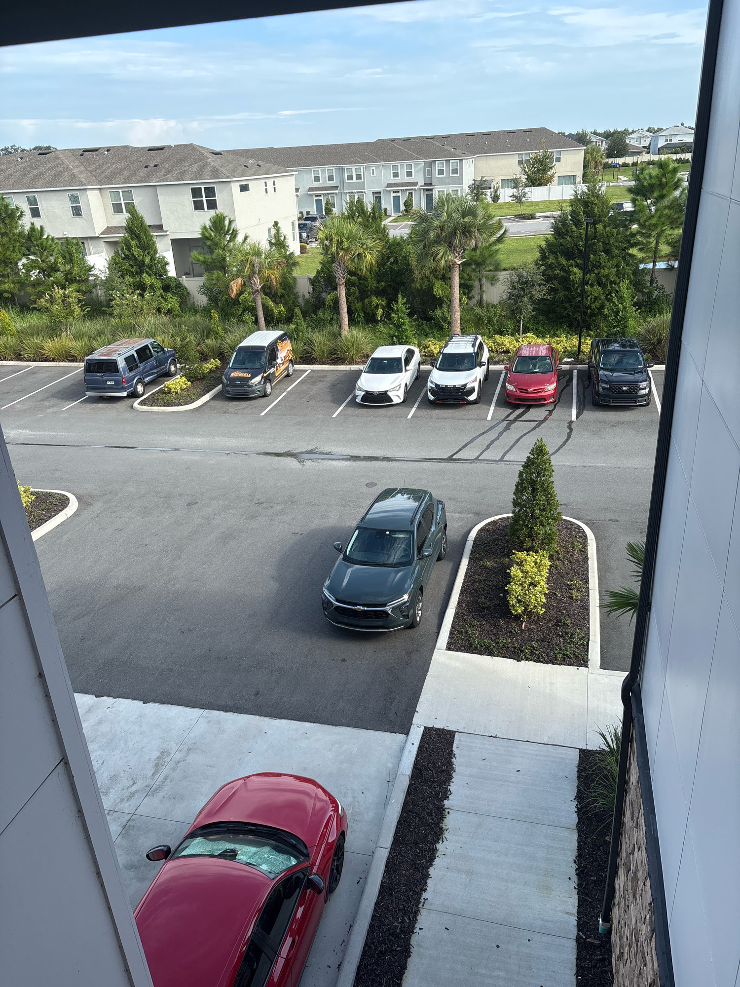 A high-angle outdoor shot shows a parking lot and residential buildings in Palmetto, United States. The parking lot is mostly empty, with a few cars parked including a dark gray SUV, a red sedan, and a black SUV. A red sports car is parked in a driveway in the foreground. In the background, a row of townhouses is visible, with a few trees and palm trees scattered throughout the landscape. The sky is partly cloudy.