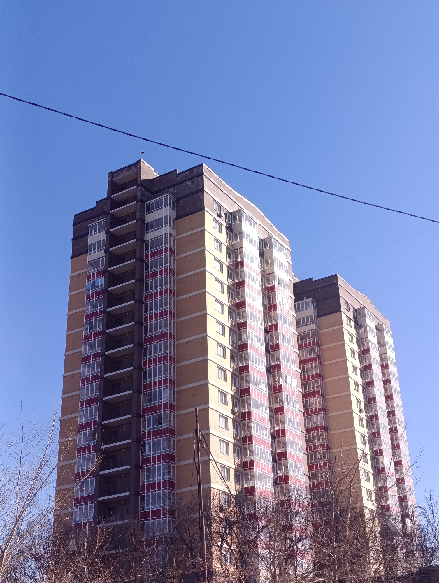 A low-angle shot reveals a tall, modern apartment building against a clear blue sky. The building appears to be located in Krasnoyarsk, Russia. The building's facade features a combination of light-colored panels, possibly tan or beige, with sections of windows framed by a red or reddish-brown material. Balconies can be seen on one section of the building. The building has a darker top section with a flat roof. Several bare trees are visible in the foreground, indicating it may be late fall or winter. A utility pole and a power line cut across the frame.