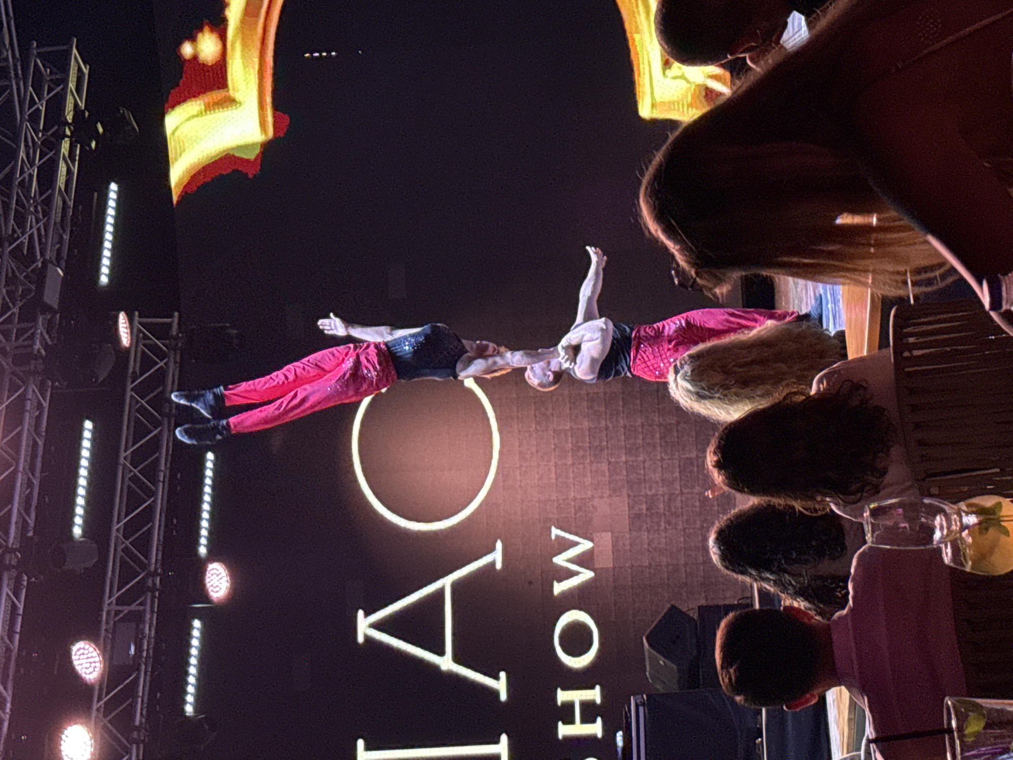 A live performance is taking place on a stage in Bodrum, Turkey, with two acrobats in vibrant red and black outfits balancing on each other. The upper acrobat is performing a handstand on the shoulders of the lower acrobat, who is standing upright. Behind them, a large screen displays illuminated text that reads "DIAC SHOW." The stage is equipped with lighting rigs and speakers. In the foreground, the audience is seated, with their backs to the camera, enjoying the show. Drinks are visible on tables in front of some audience members. The overall atmosphere suggests an evening entertainment event.