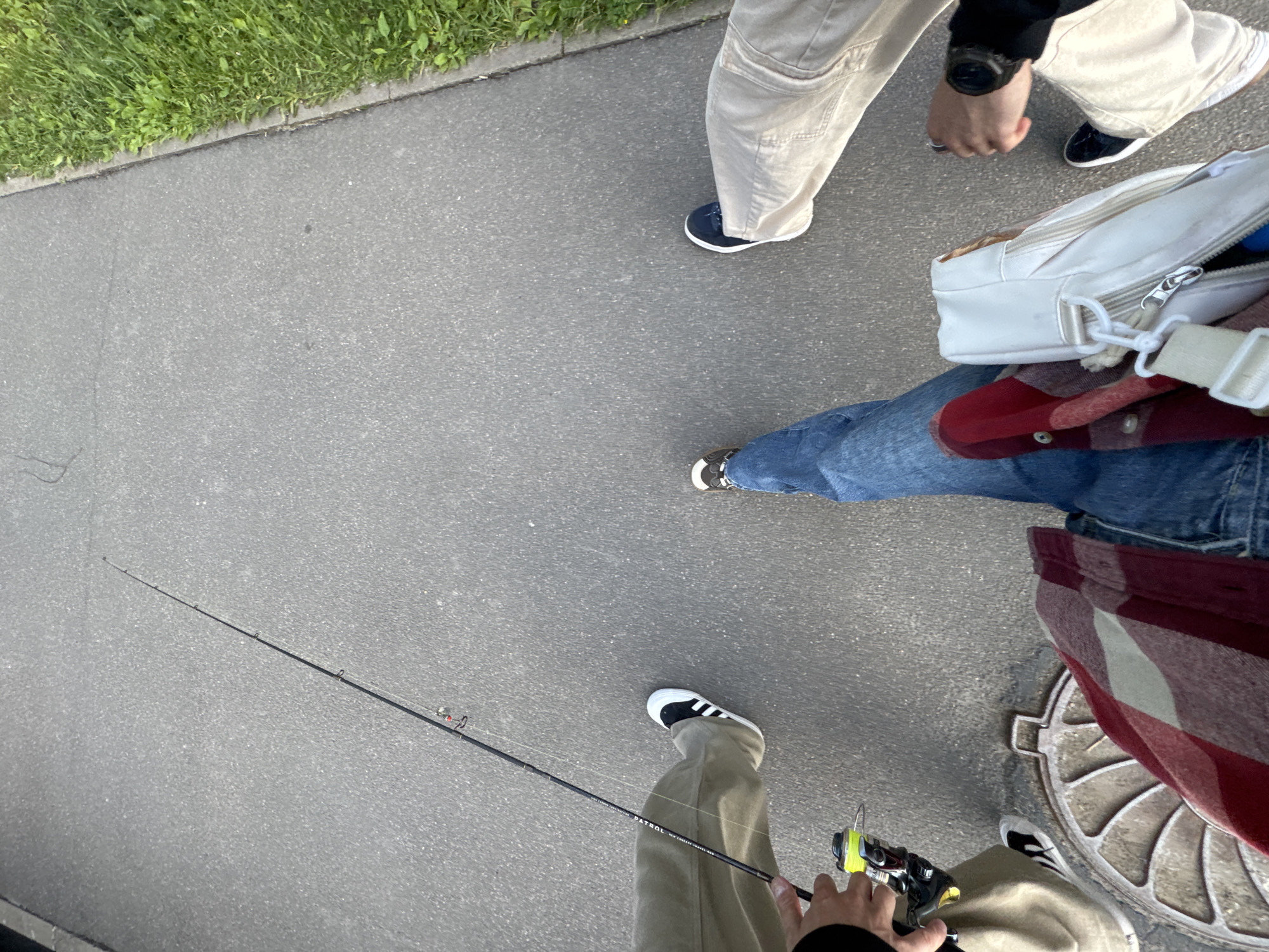 The image depicts a low-angle, POV shot of three individuals walking on a paved path adjacent to a grassy area. The path appears to be asphalt with some cracks and is next to a concrete curb. The person in the foreground, presumably the photographer, is wearing blue jeans and a red and white checkered shirt, carrying a white crossbody bag. Their left hand is holding a fishing rod with a yellow reel. To the left, another person is wearing light brown pants and black and white sneakers, also holding a fishing rod. To the right, a third person is wearing light brown pants and navy blue sneakers. The environment suggests an outdoor urban or park setting. The image is taken during daylight.