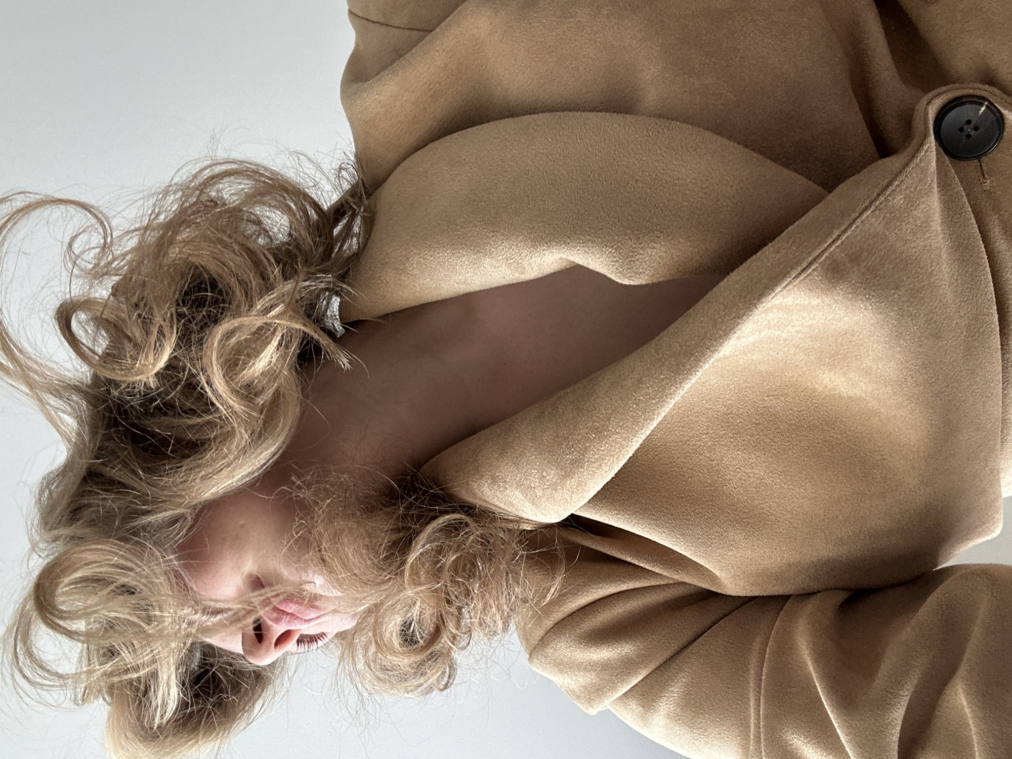 A low-angle shot shows a fair-skinned person with wavy blonde hair. Part of their face, including their closed eyes, nose, and lips, is visible. They are wearing a tan-colored coat with a button visible in the upper right corner. The background is a light grey or white. There are no indications of a location in the city of Kazan, Russia, or any other specific place.