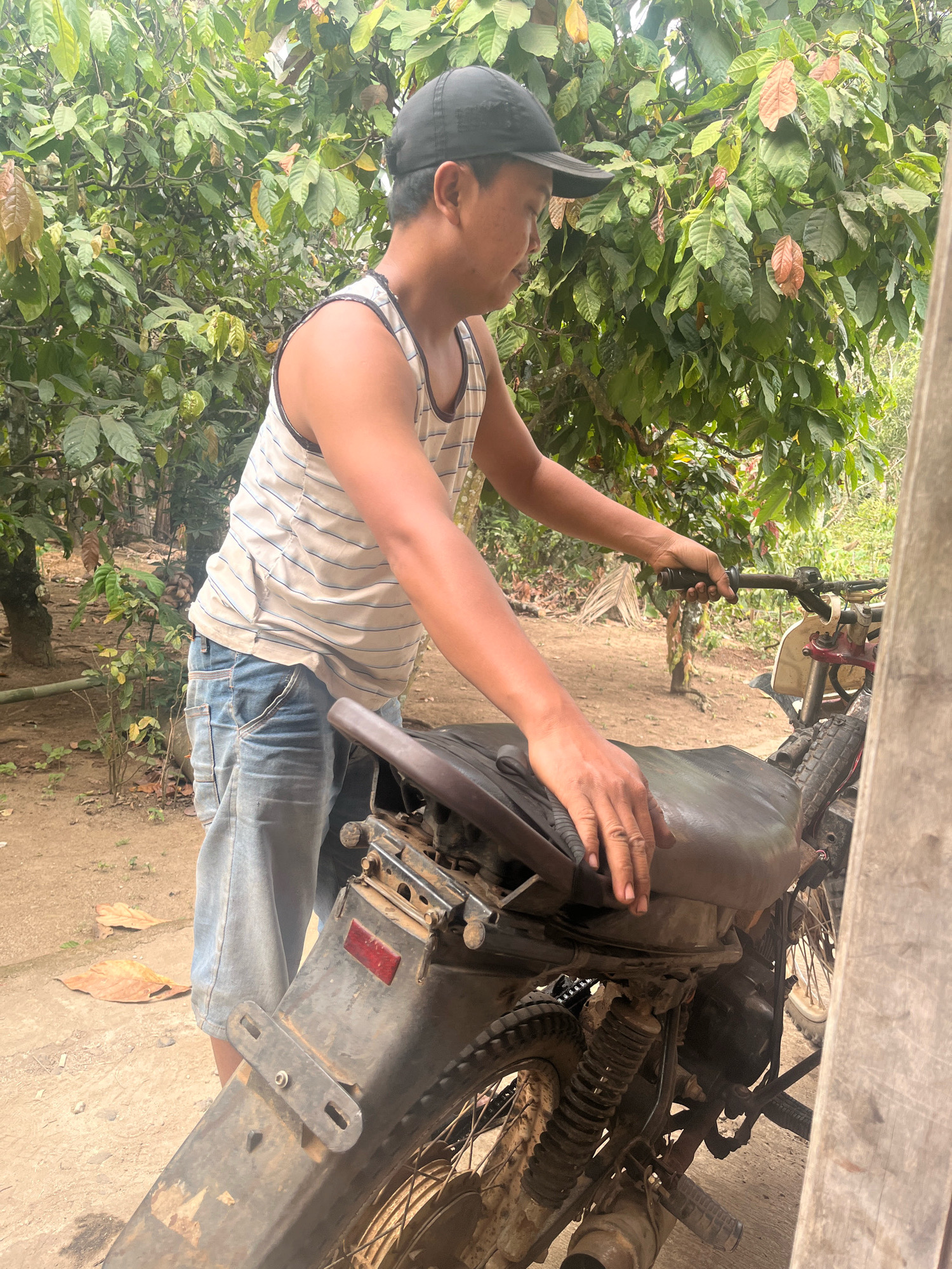 A man wearing a black baseball cap, a striped tank top, and denim shorts is standing next to a dirt bike in an outdoor, rural setting. The man is looking down and appears to be interacting with the motorcycle, with his left hand resting on the seat and his right hand on the handlebars. The motorcycle is dark colored and appears to be well-used. The background consists of lush green foliage, suggesting a tropical environment. The ground is covered in dirt and fallen leaves. The scene appears to be in the city of Kebumen, Indonesia, a region known for its agricultural landscape and rural communities.