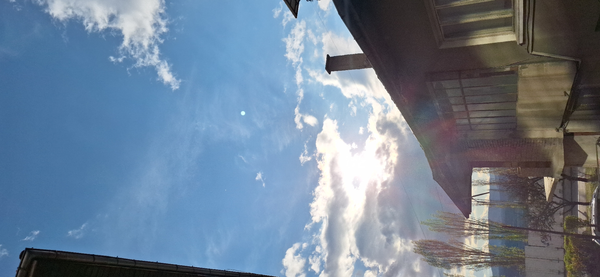 The image depicts a view looking up at the sky in Blagoevgrad, Bulgaria. The sky is primarily blue with scattered white clouds and the sun visible through a break in the cloud cover. Buildings are partially visible along the top and bottom edges of the frame. A chimney extends from the roof of one building. Trees are visible near the building on the right side.