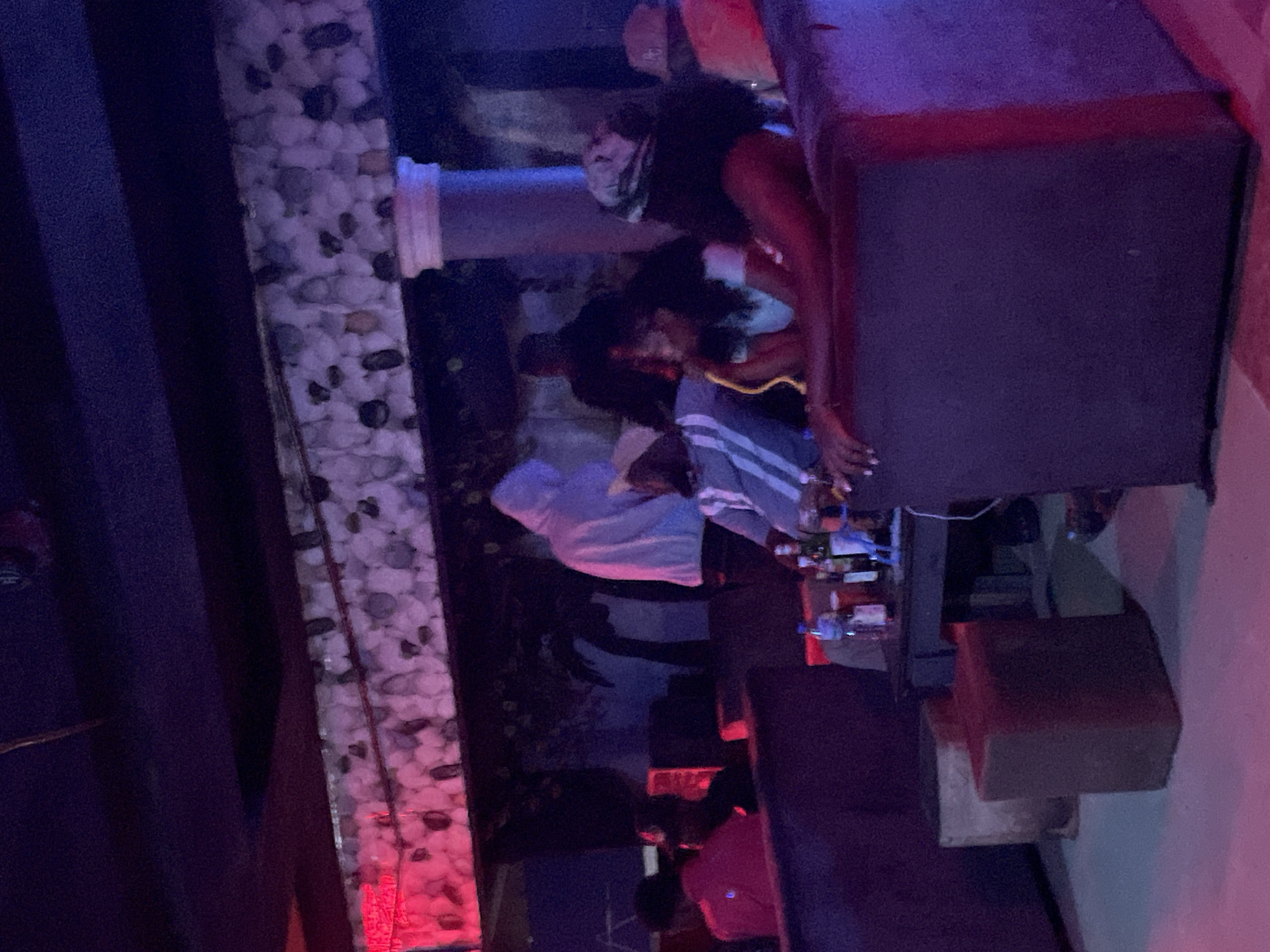 The image depicts an indoor scene, likely a lounge or club, in Durumi, Nigeria. The lighting is dim and atmospheric, with prominent red and purple hues, suggesting nighttime activity. In the foreground, two individuals are seated on a plush, dark-colored couch. One, a woman with dark curly hair, is leaning back, her arm resting on the couch's armrest. She is wearing a light-colored top and has a headscarf or similar covering. To her left, another person, possibly a woman, is also seated, appearing to be engaged in conversation or activity. Their face is partially obscured.
In the middle ground, a table holds several bottles of alcohol and drinks, indicating a social gathering. Another person in a light-colored hoodie is visible behind the table, seemingly observing. Further back, other figures are blurred and indistinct, contributing to the overall ambiance of a crowded, dimly lit space. The background features a textured wall with what appears to be decorative stones and hints of foliage, possibly an artificial mural. The presence of various seating arrangements, tables, and the overall setup suggest this is a place for leisure and entertainment within the Durumi district.