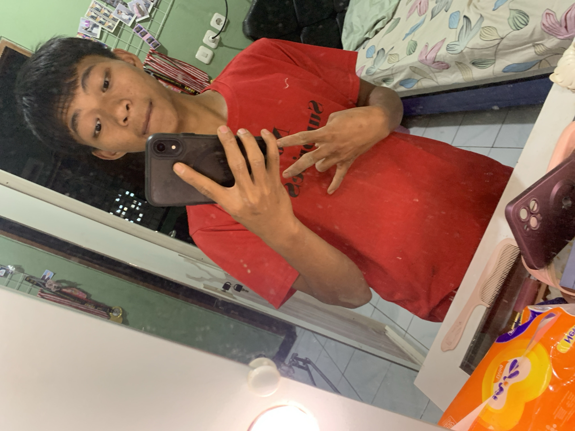The image is a mirror selfie taken by a young male indoors. He is wearing a red t-shirt and is holding a black smartphone in his left hand, taking the picture. His right hand is held up, with two fingers extended in a peace sign. The background shows a bedroom with a bed covered in floral-patterned sheets. On the wall behind him, there are several photos and what appear to be small decorative items. To the left of the frame, a vanity mirror with a lightbulb is visible, reflecting a portion of the room. A white countertop with items like a comb and a package of tissues is in the foreground. The lighting suggests it is likely daytime or the room is well-lit. The exact location within Tanggung, Indonesia, cannot be determined from the image alone.
