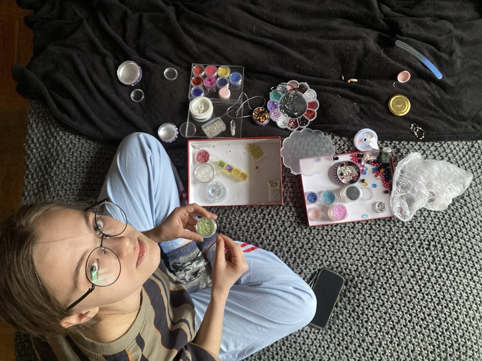 A young person with glasses sits cross-legged on a gray textured blanket. The person is wearing a striped shirt and blue sweatpants and is holding a small container of green beads. Around them are various containers and boxes filled with beads and crafting supplies. Items include clear plastic containers with colored beads, a flower-shaped organizer, a box with different beads, and various loose beads. There is also a black phone lying on the blanket. The scene is illuminated with natural light, with a black cloth as background. Moscow, Russia, cannot be determined from the image.