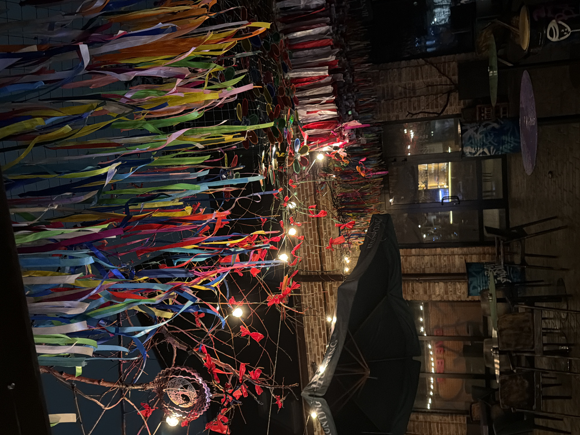 The image shows an outdoor patio or dining area, possibly at a restaurant or bar, at night. The location is Rostov-on-Don, Russia. The area is decorated with colorful ribbons suspended overhead, creating a festive atmosphere. There are also lit branches, possibly decorative trees or art installations, with red ribbons or ornaments, also with light bulbs, entwined in them.
In the background, there is a brick building with windows, and through them, there appears to be an interior space with additional lighting. An outdoor umbrella is also visible. Chairs and tables are set up in the patio area, suggesting a seating arrangement for customers. There are no people visible in the image.
