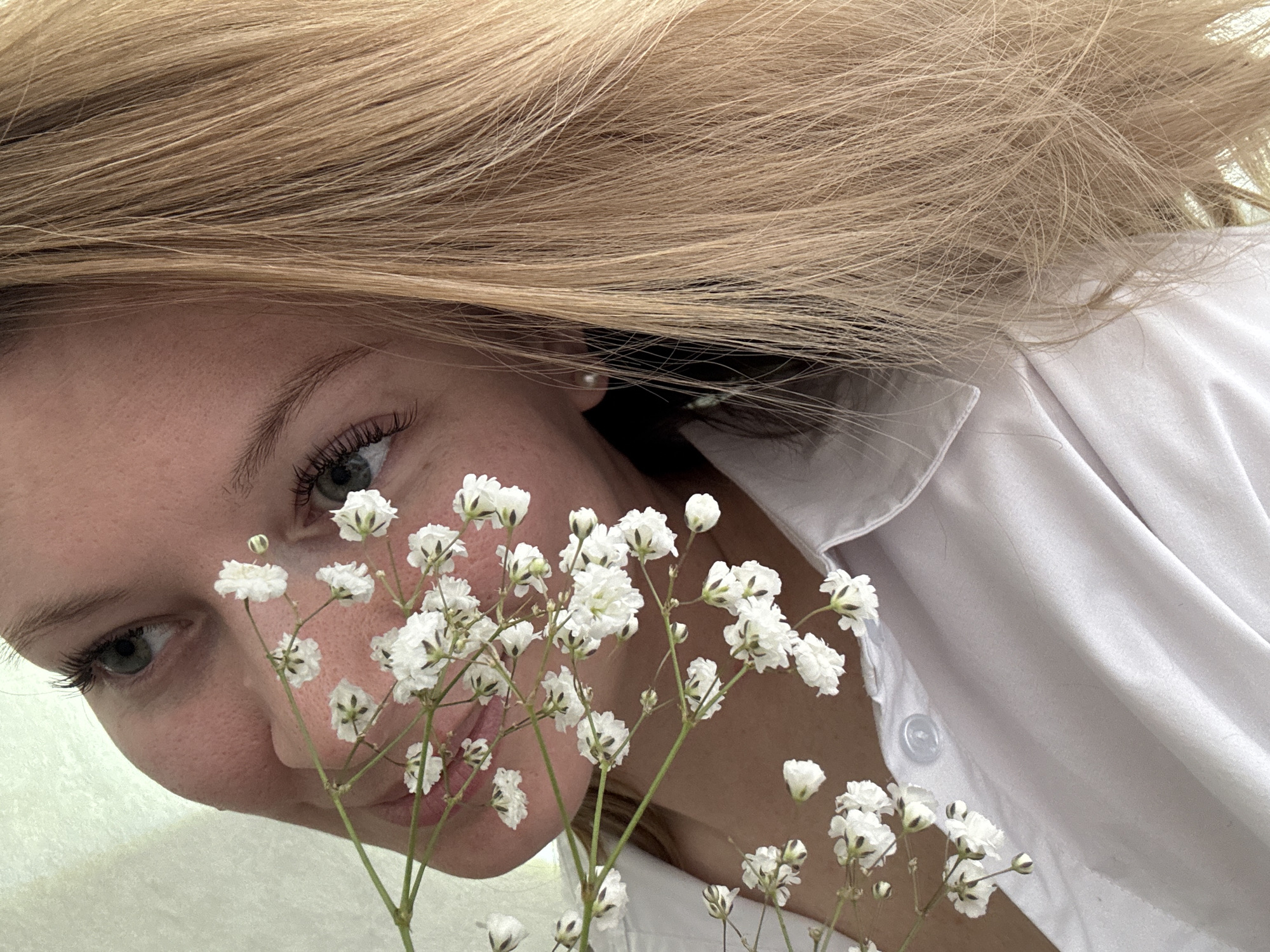 A woman with blonde hair is shown from a low angle, close-up perspective. She is holding a small bouquet of baby's breath flowers near her face. She is wearing a white button-down shirt. The background is out of focus. No information about the location in Chaikovsky, Russia can be determined from this image.