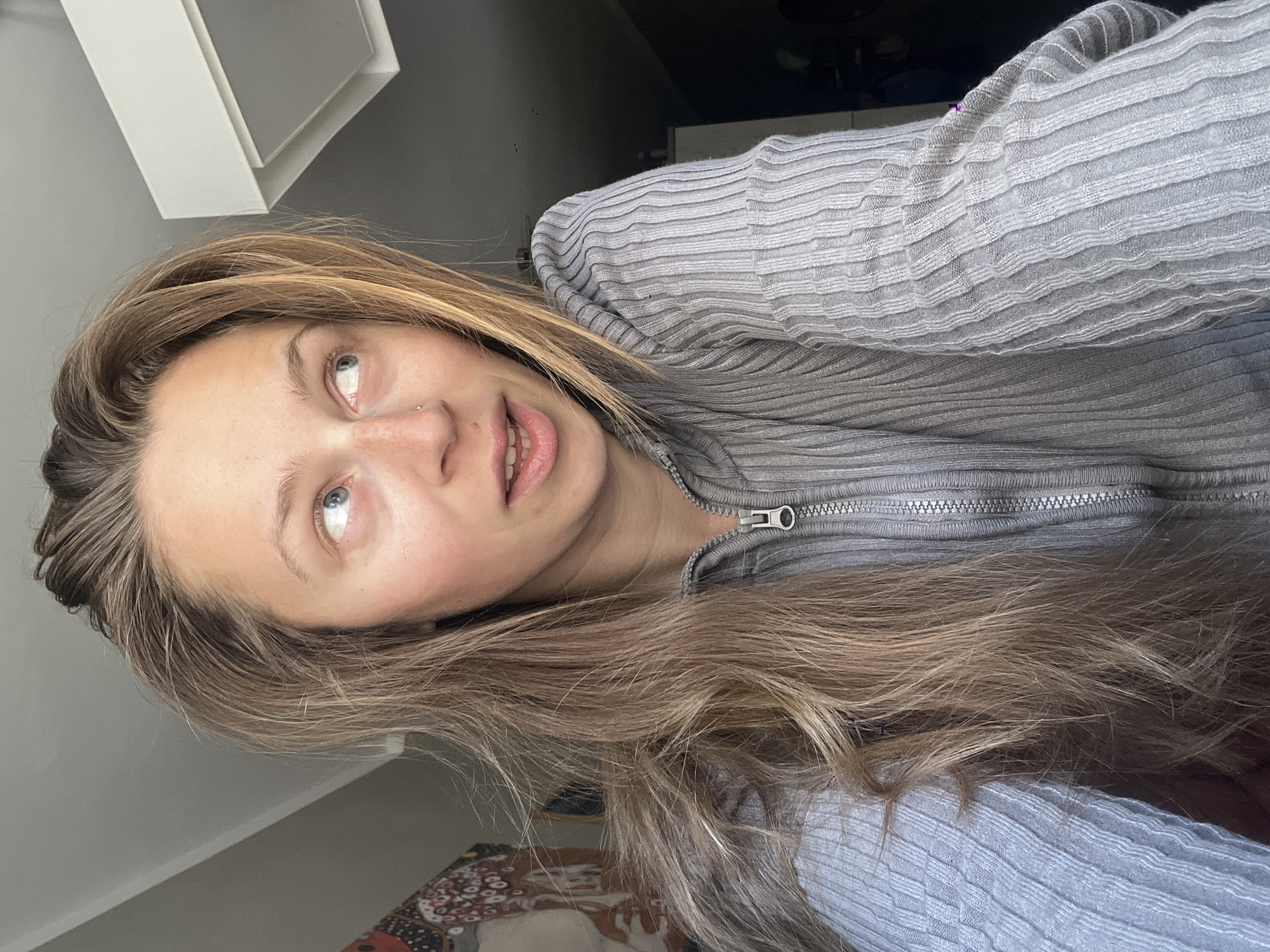 A young woman with long brown and blonde hair is wearing a gray zippered sweater and looking upwards and to the left of the frame. She appears to be indoors, possibly in a residential setting based on the decor. The location in Bečići, Montenegro, is not discernable from the image provided.