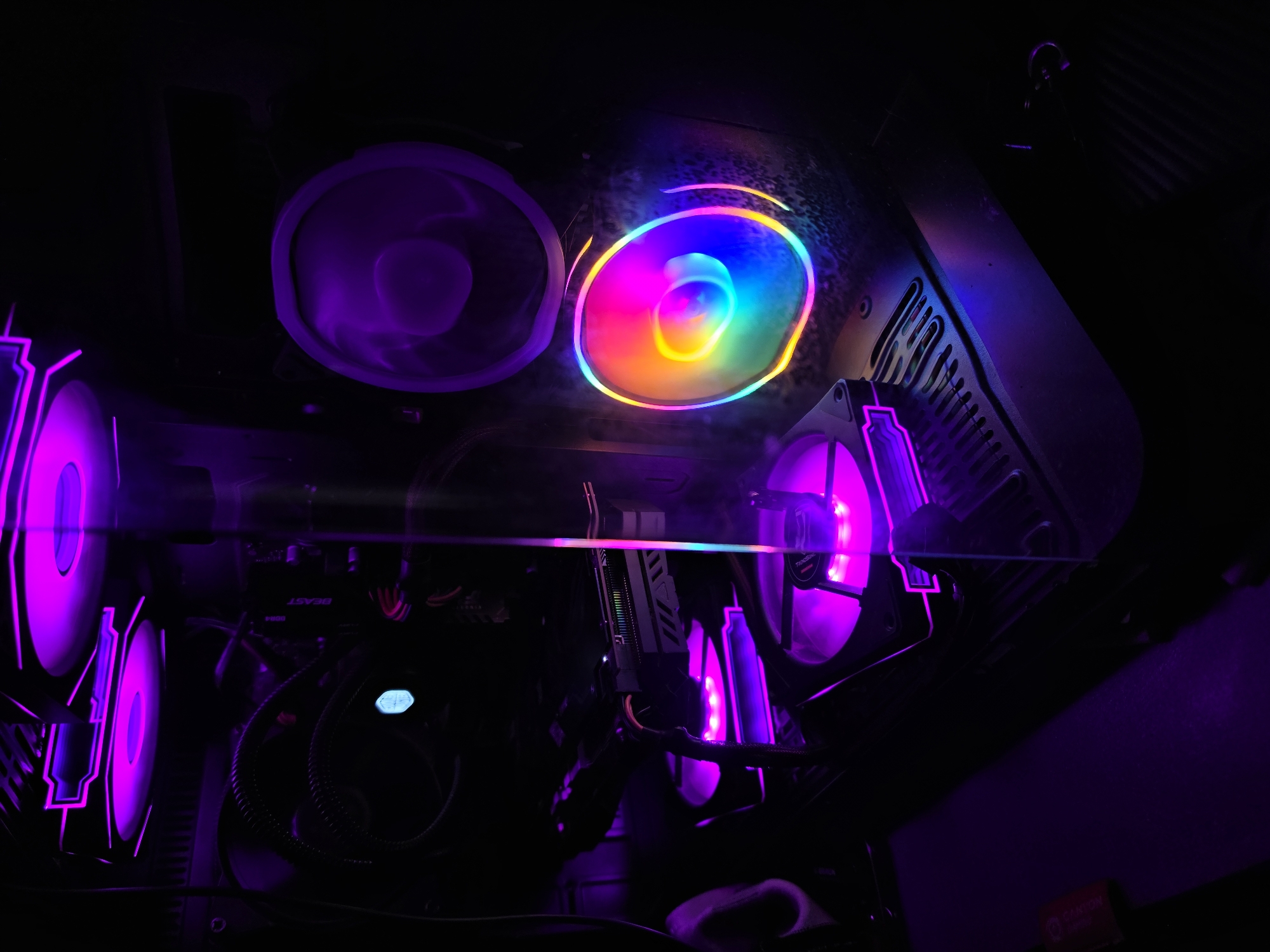 The image shows the interior of a computer with multiple fans illuminated by purple and rainbow-colored LEDs. The components are mostly in darkness, with the lighting from the fans creating streaks and reflections on the case. One fan in the lower right displays a swirling rainbow of colors, while others are lit with a solid purple hue. A graphics card is visible mounted on the motherboard. The computer appears to be situated in a dimly lit environment, with a dark surface in the foreground. There are no people visible in the image, and no contextual details suggest a specific location in Aleksandrovo, Bulgaria.