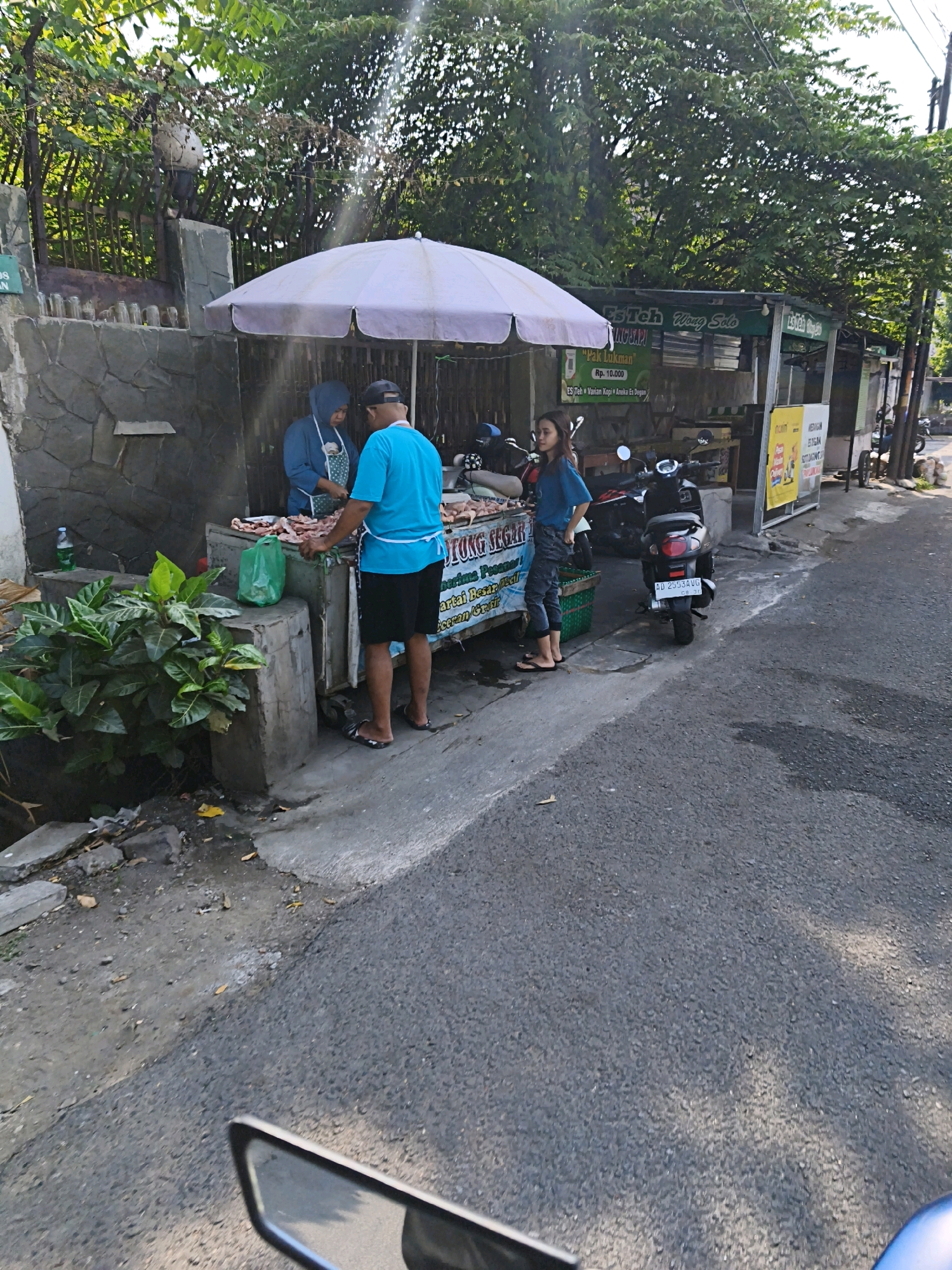 The image shows a street-side stall in Surakarta, Indonesia, selling raw meat, likely chicken. A woman in a blue hijab and apron stands behind the stall, and a man in a blue t-shirt and black shorts is preparing the meat. A young woman in a blue t-shirt and grey pants stands to the side, and two motorcycles are parked nearby. The stall is covered by a large, light-colored umbrella. Signs on the stall indicate it is selling "Daging Segar" (fresh meat) and other food items. The scene is set against a backdrop of trees and a stone wall, with a paved road in the foreground. The overall atmosphere suggests a typical day at a local market or food stall in an urban Indonesian setting.