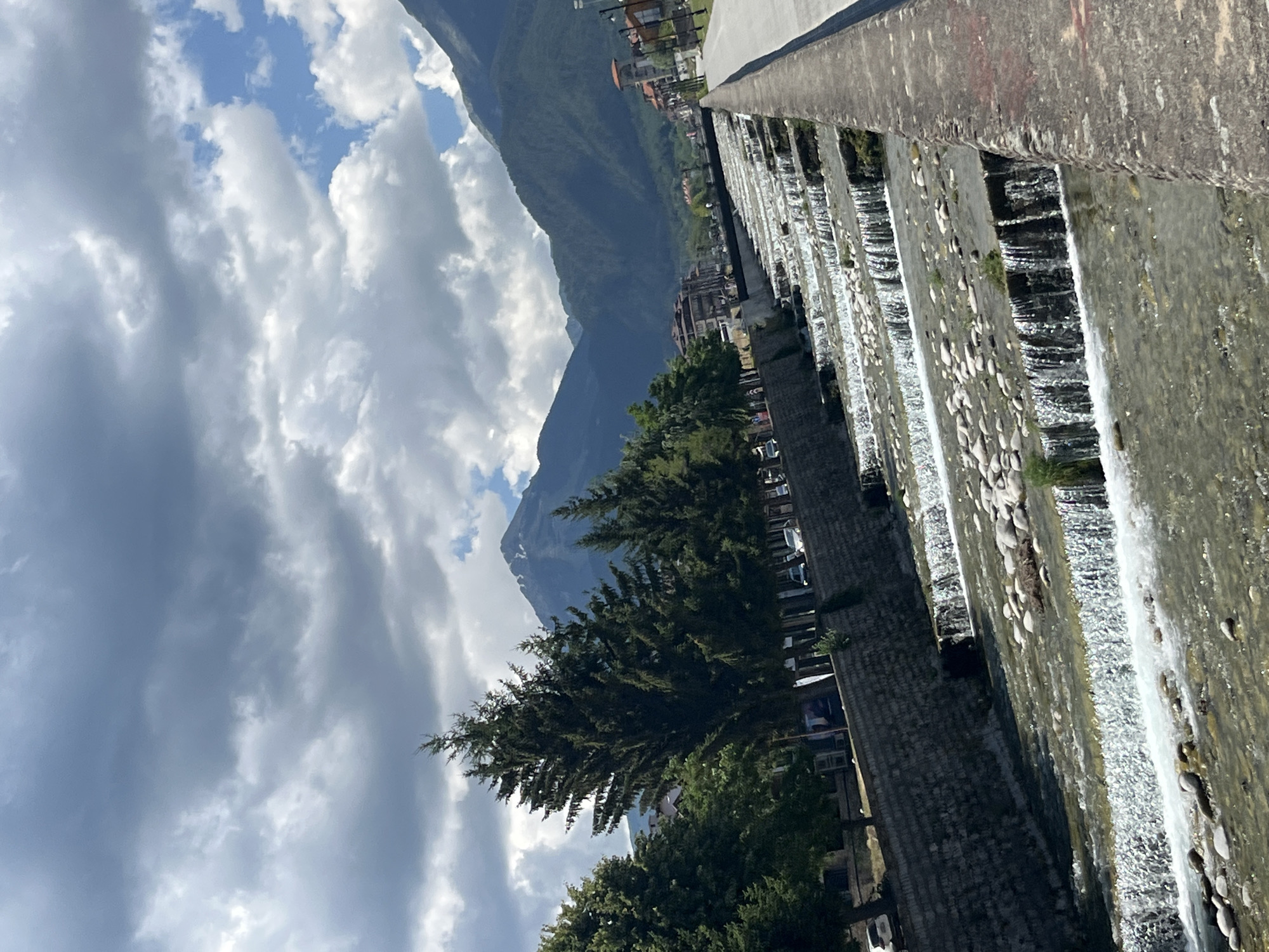 This is a landscape photograph taken in Bansko, Bulgaria, featuring a river with a series of man-made cascades flowing through a town. The river, with its clear water and stony bed, runs parallel to a paved walkway on the right and a stone wall retaining an elevated area with trees and buildings on the left. The cascades are constructed with stone or concrete, creating multiple levels of flowing water.
In the background, lush green trees line the riverbank, behind which lie residential and commercial buildings with red-tiled roofs. Further in the distance, imposing mountains with forested slopes rise dramatically, with a snow-capped peak visible in the center. The sky is filled with a mix of dramatic, dark storm clouds and patches of bright blue sky. Several cars are visible parked or driving along the road on the left side of the river. No people are clearly discernible in the foreground or mid-ground, contributing to a sense of quietude. The overall atmosphere is one of natural beauty juxtaposed with urban development.
