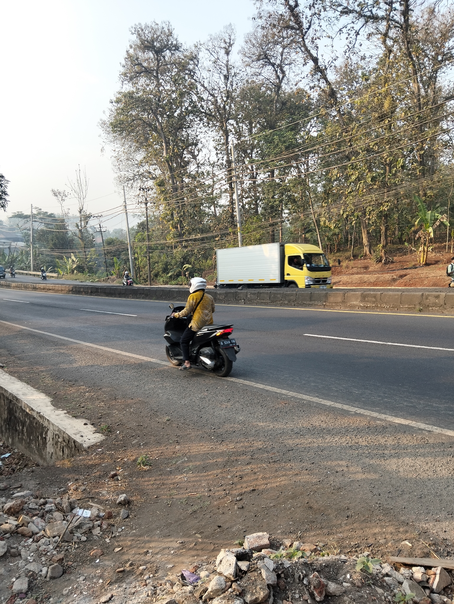 In Durenombo, Indonesia, a street scene unfolds under a hazy sky. The focus is on a person riding a black motorcycle, wearing a yellow long-sleeved shirt and a white helmet, moving from left to right. The motorcycle is in the foreground, on the right side of the asphalt road. In the background, a yellow truck with a white cargo box is also traveling from left to right. Further in the distance, several other motorcycles are visible on the road, and a line of trees borders the left side of the scene. The environment appears to be rural or semi-rural, with a dirt shoulder on the right side of the road littered with rocks and debris, and power lines stretching across the frame.