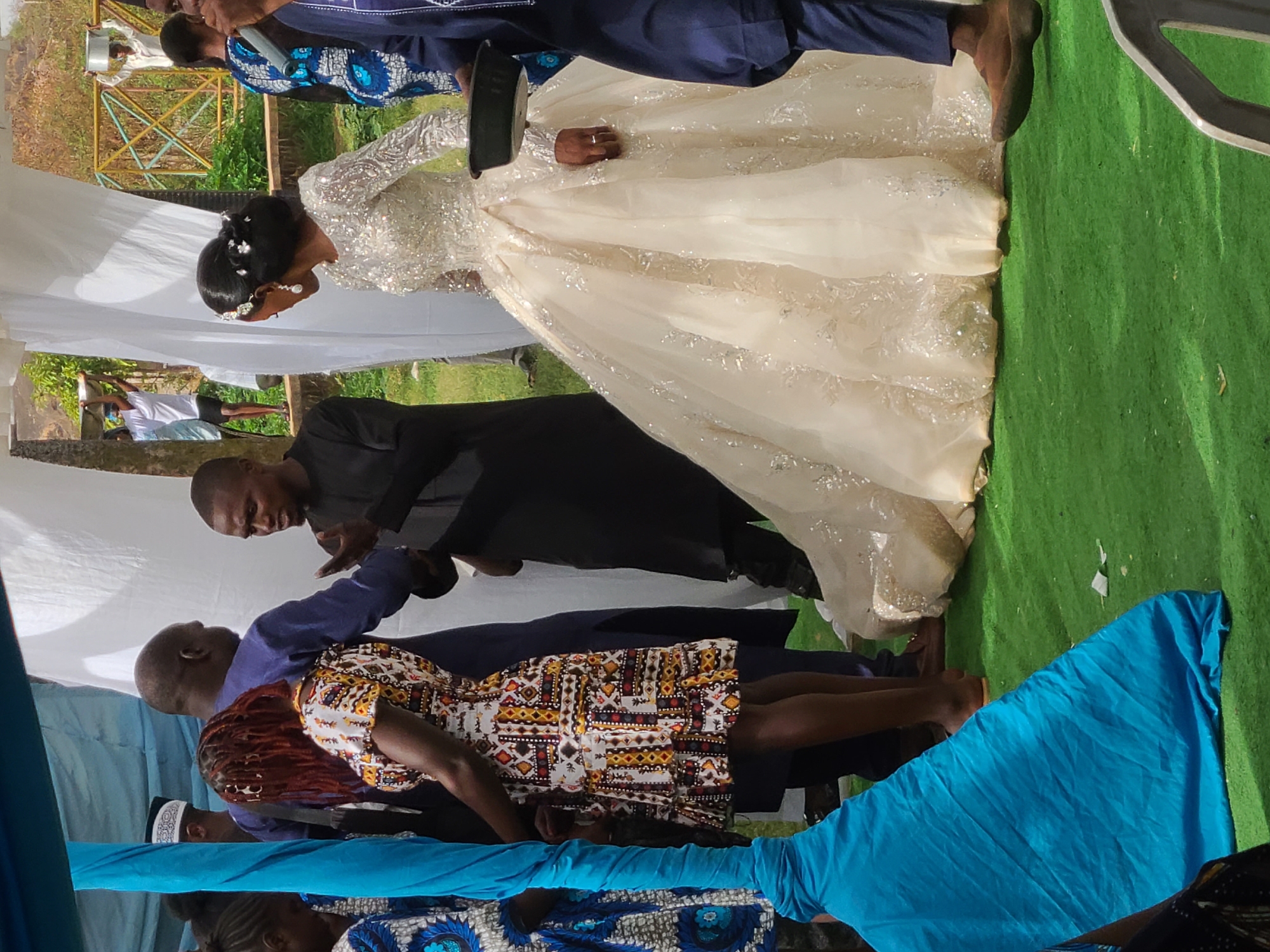 The image captures a wedding ceremony in Jos, Nigeria. A bride in an elaborate, light-colored gown with long, possibly embellished sleeves stands on green turf, seemingly being attended to by a man in a black suit who is crouched down. The bride's head is bowed, and she wears a headpiece. Several other individuals are present, dressed in a variety of colorful attire, including patterned fabrics and blue suits. The scene is partially framed by a structure draped with white and blue fabrics. A glimpse of greenery and a distant metal structure is visible in the background.