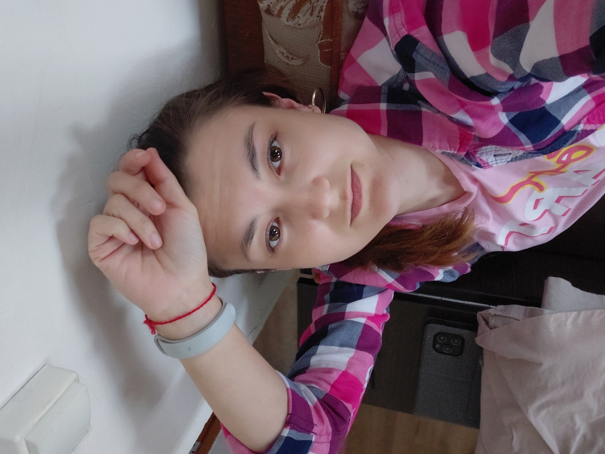 A woman is lying down and looking at the camera with her right hand on her forehead. She is wearing a pink and blue plaid shirt over a pink t-shirt. A grey fitness tracker is visible on her left wrist, along with a red string bracelet. A gold hoop earring is worn on her right ear. In the background, a dark object, possibly a tablet or phone, is visible, as well as a pillow with a pattern. The lighting suggests an indoor environment, possibly a room. There is no indication of the location being Tashkazgan, Russia.