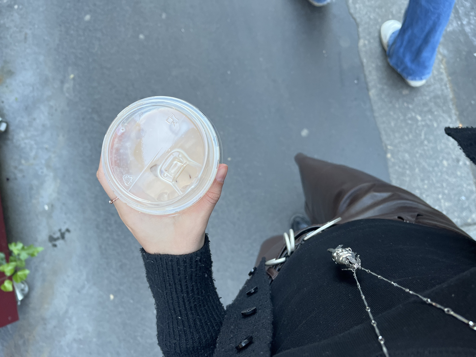 An overhead shot captures a person holding a clear plastic cup with a light-colored beverage, likely coffee or tea with milk, while standing or walking outdoors on what appears to be a concrete or asphalt surface. The person's left hand, adorned with a small ring on one finger, holds the cup. The cup's lid shows a "20" marking, a recycling symbol with "PP" inside, and an icon resembling a dishwashing symbol.
The person is dressed in a black long-sleeved sweater or cardigan with black buttons running down a front placket. A silver chain necklace with an intricate, possibly rose-shaped, pendant is visible against dark pants or a skirt. A brown, possibly leather, object (perhaps a bag strap or part of a coat/boot) can be seen next to their body.
In the background, on the right side of the frame, another person's lower legs and feet are visible, wearing blue jeans and white shoes. On the far left, a small green plant is partially seen within a reddish planter. The ground is a textured gray, indicating an urban outdoor setting, possibly a street or sidewalk. The lighting suggests it is daytime, with no harsh shadows. Based on the provided context, the scene takes place in Paris, France.