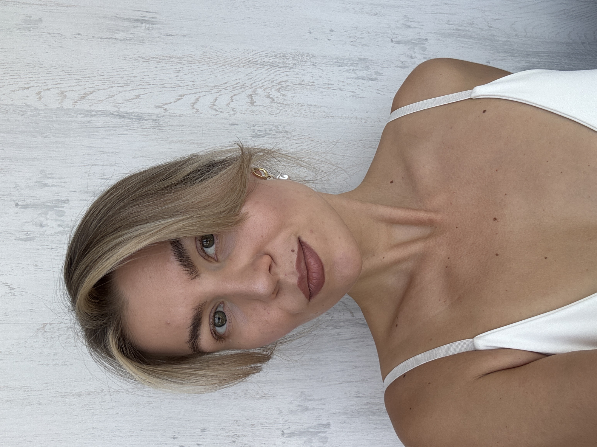 A rotated eye-level close-up captures a woman with fair skin, light blonde hair styled with dark roots and lighter highlights, and green eyes. She has well-defined eyebrows, full lips with a neutral lip color, and a small mole visible near her lip. The woman is wearing a white top with thin straps, revealing her shoulders and collarbone. The background appears to be a white, wood-grain surface. Her pose is canted, causing her head to tilt towards her left shoulder. A piece of jewelry is visible in her left ear. The location appears to be indoors.