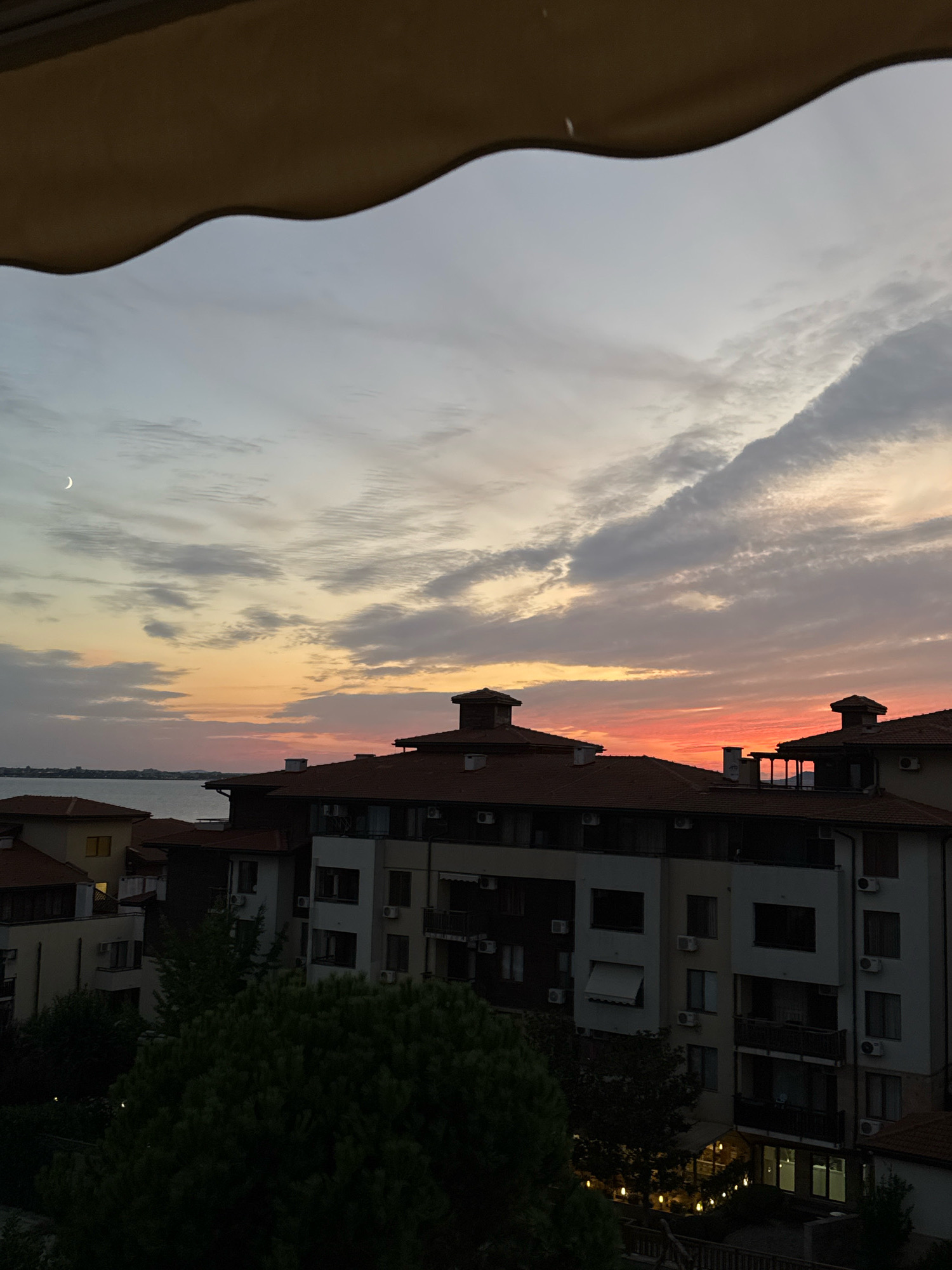 The image captures a sunset scene over a coastal city, likely Sveti Vlas, Bulgaria, given the architectural style of the buildings which appear to be apartment complexes and residential structures with tiled roofs. The sky displays a gradient of colors, transitioning from a pale blue at the top to hues of orange, pink, and red near the horizon, indicative of a sunset. Wispy clouds are scattered across the sky. In the upper left quadrant, a crescent moon is visible. Below the sky, several multi-story buildings are prominent, featuring balconies, windows, and air conditioning units. Some of these buildings have red-tiled roofs, a common architectural feature in the region. In the foreground, a dark silhouette of evergreen trees obscures the lower parts of the buildings. The presence of the sea is suggested by a lighter strip of water in the middle ground, separating the buildings from a distant, faint outline of a shoreline. The scene is devoid of people and immediate action. The overall impression is a tranquil evening view of a European coastal town.