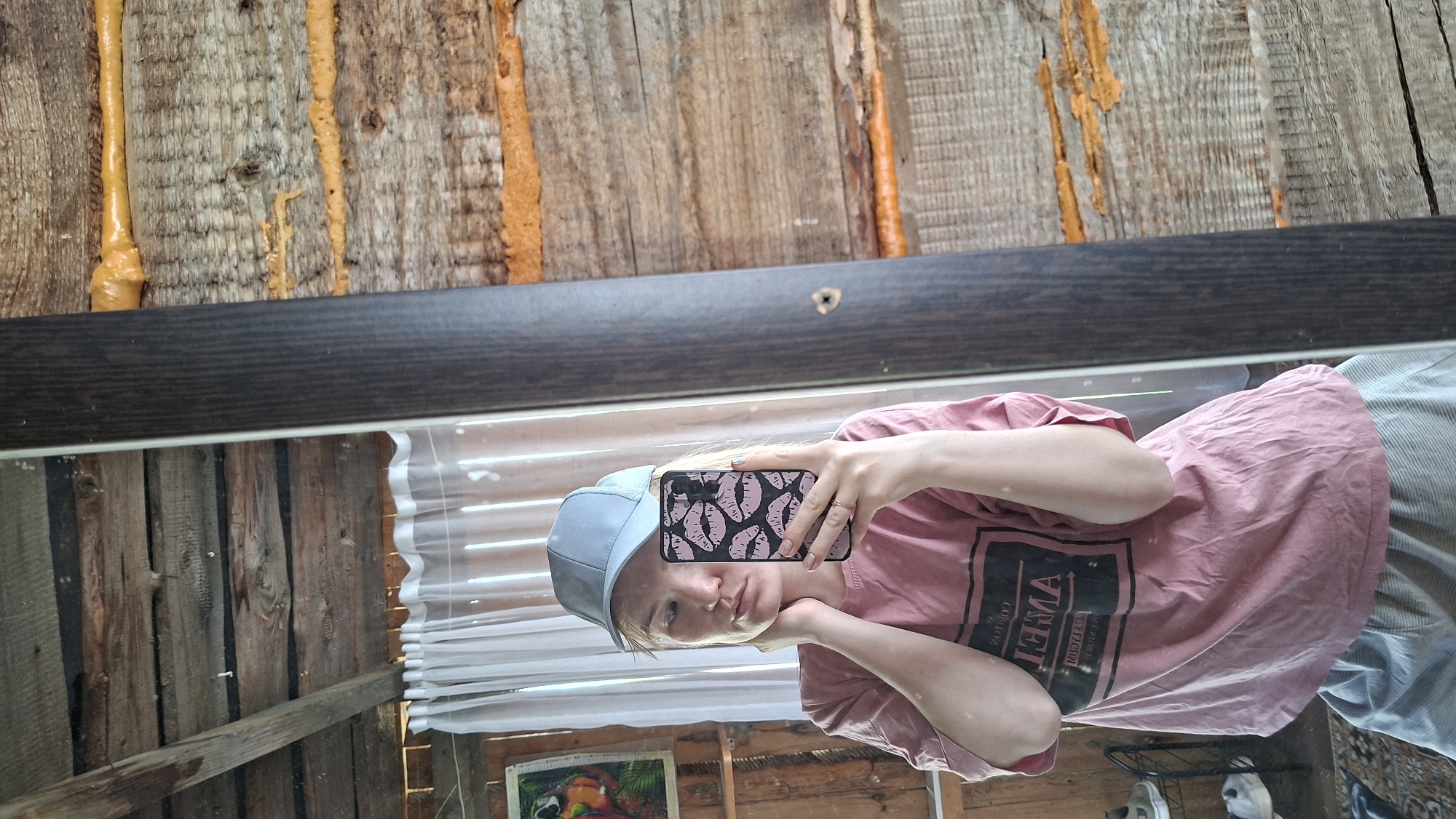 A young woman is reflected in a mirror. She is wearing a light blue baseball cap and a pink t-shirt. She is holding a phone in her hand, taking the photo. The background features wooden walls with orange sealant visible. There is a painting partially visible at the bottom of the mirror. The blinds and mirror frame are horizontal. The woman's body is angled away from the viewer. The location is Большой Букор, Russia, based on information available.