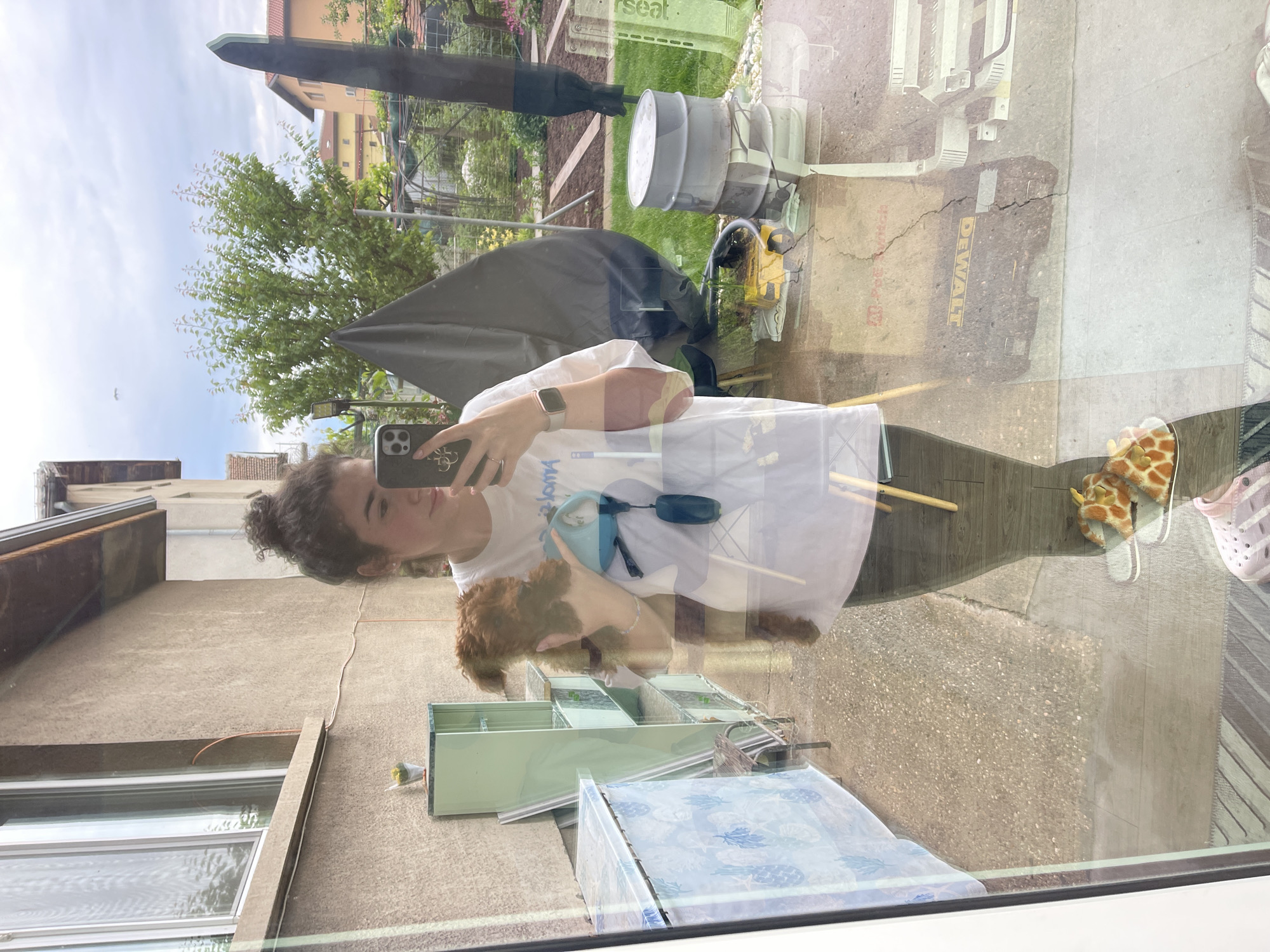 A young woman with curly hair is taking a selfie in front of a window, holding a small brown dog. The window reflects a view of a backyard with a lawn, trees, a gray garden furniture cover, a gray barrel, and some power tools. Outside the yard, buildings are visible. In the lower part of the reflection, some home repair materials, including disassembled window frames and a package of tiles are visible. Inside, near the window, there are animal-themed slippers. The photograph appears to be taken inside a house in Sofia, Bulgaria.