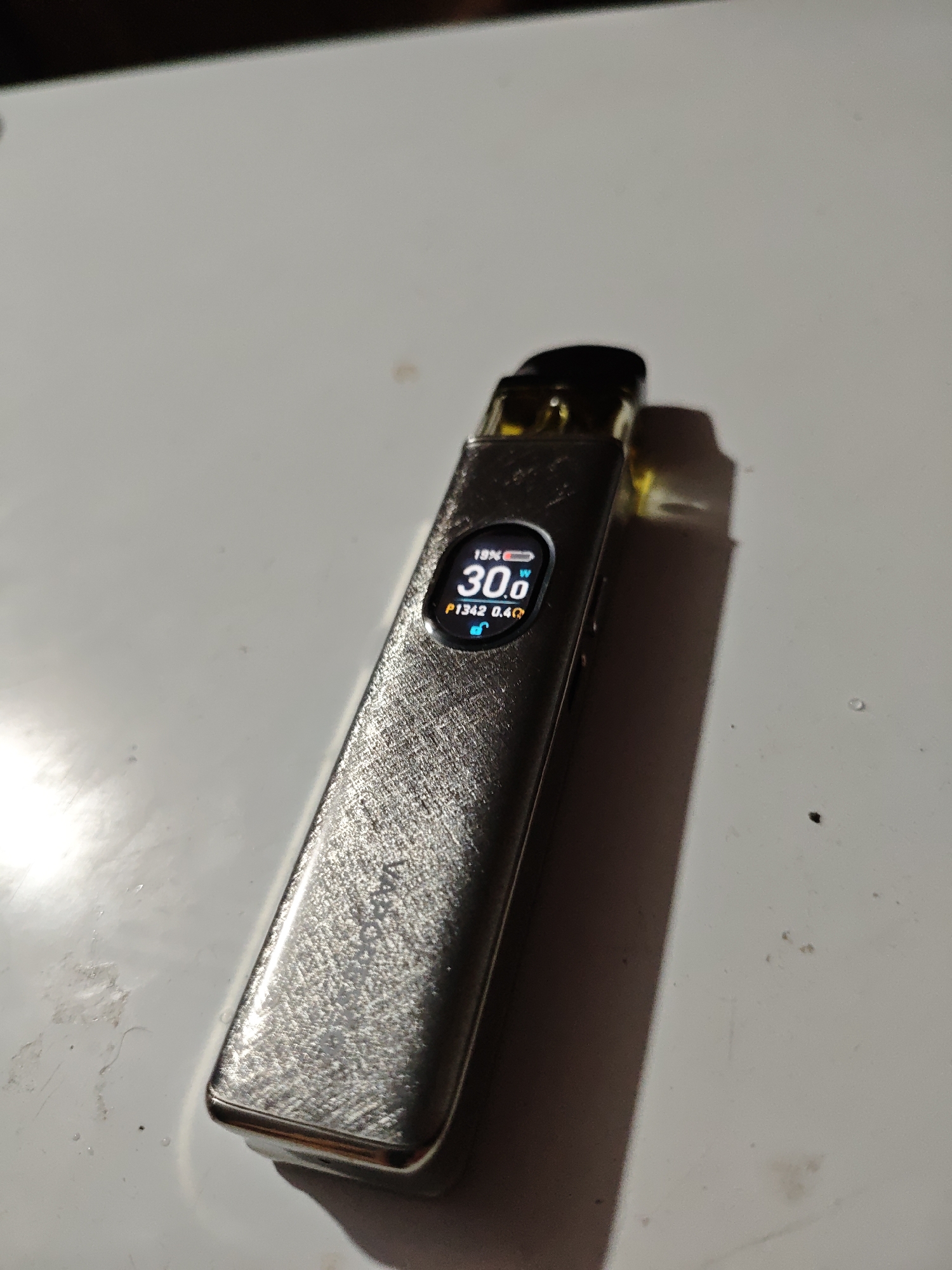 The image shows a silver colored vape pen with a screen displaying "19% ", "30.0 W", and "P1342 0.4Ω". The vape pen is lying on a white surface, likely a table, with a shadow cast to its right. There are a few small dark specks on the white surface, possibly dirt or debris. The background is dark and out of focus. The location in the city of Akhmetovskaya, Russia is not depicted in the image.