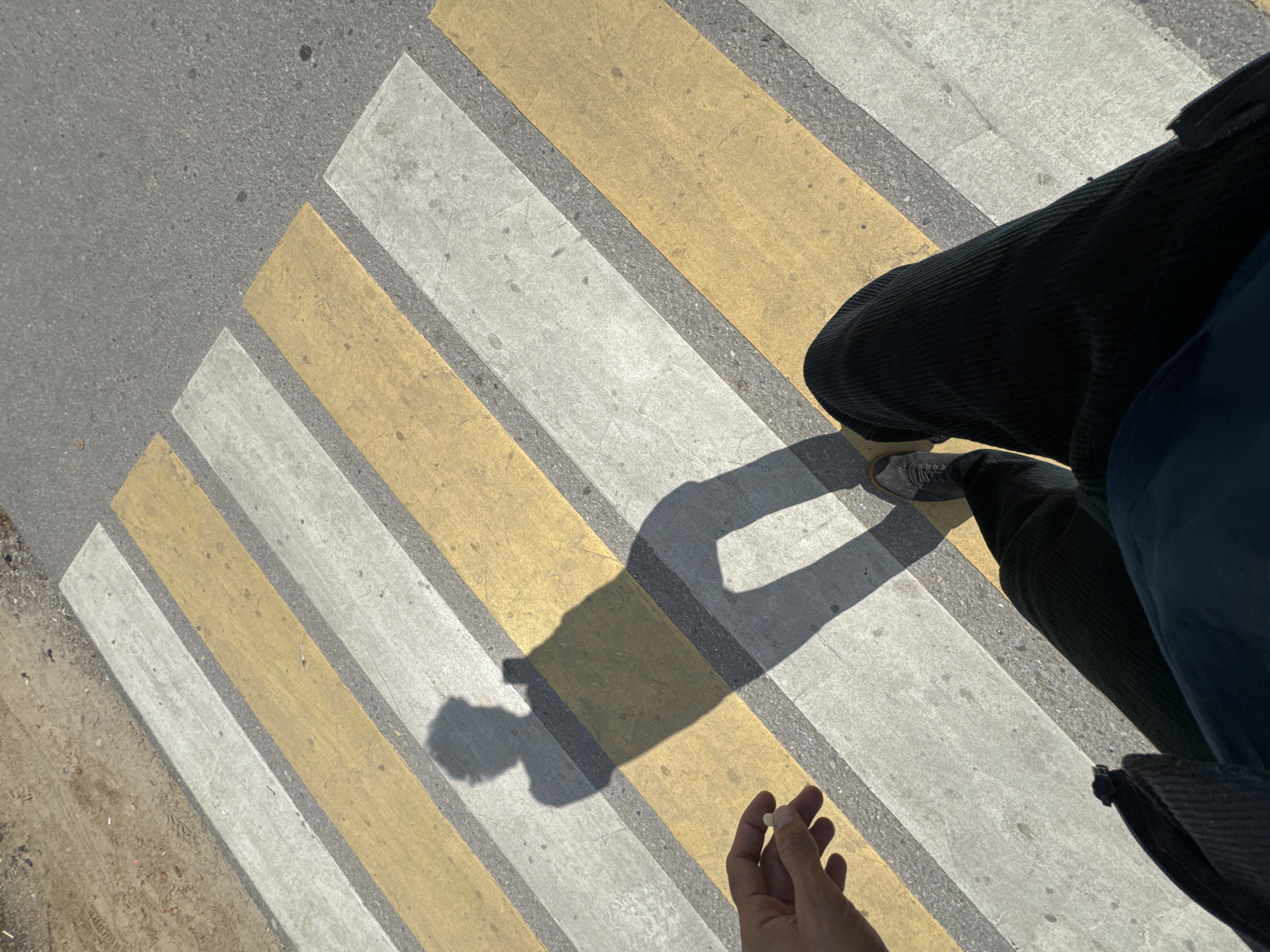 A first-person perspective shot shows a person standing on a pedestrian crossing. The crossing is marked with alternating yellow and white stripes. The person's shadow is visible on the crossing, cast by the sunlight, and it shows the shape of their body holding a smartphone. The person is wearing dark corduroy pants and dark shoes. A hand is visible in the lower left of the frame, holding a small white object, possibly a pill. The background shows the asphalt road and some dirt on the left side. The location is given as Unknown, Unknown.