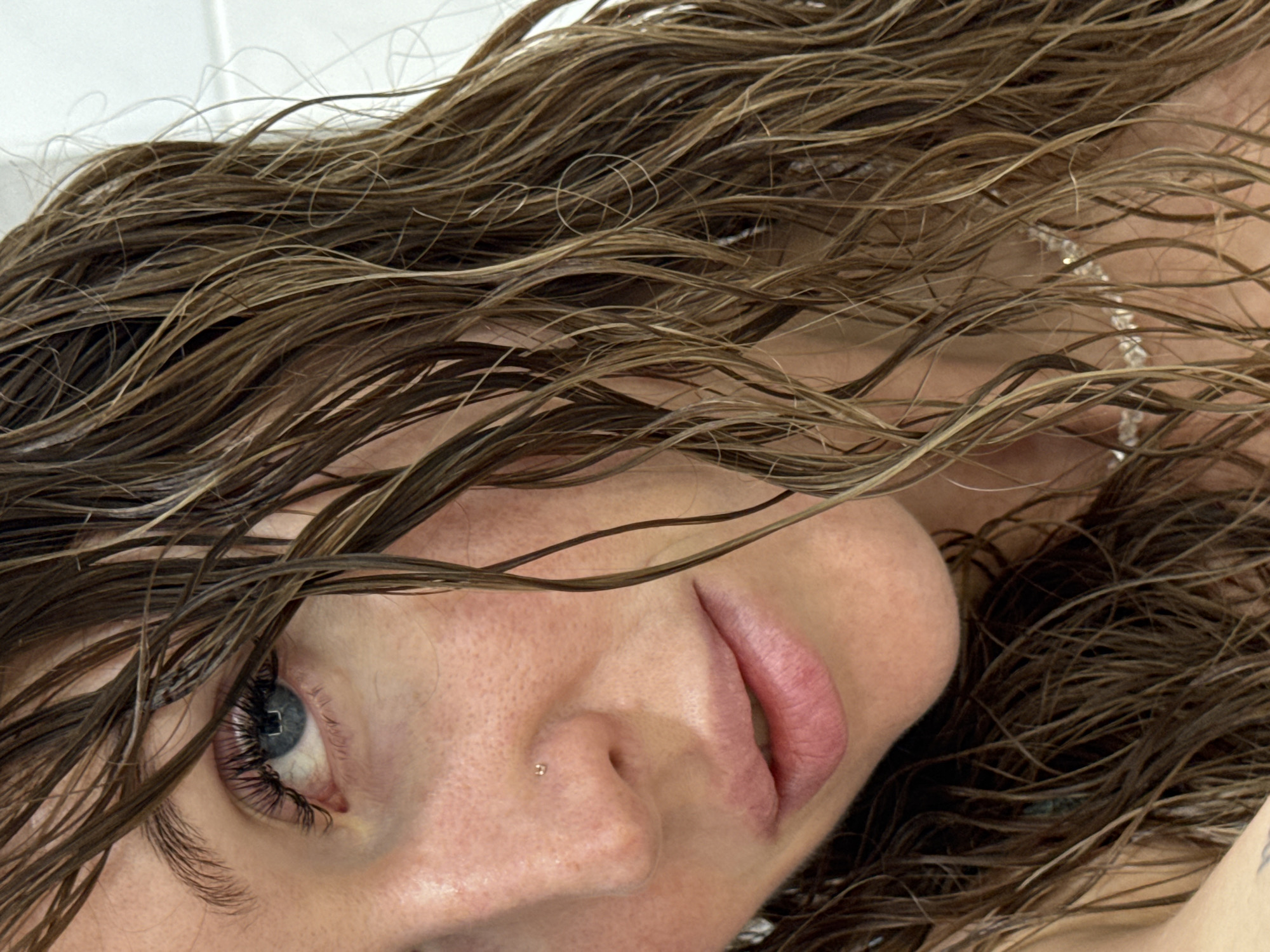 A close-up, headshot of a woman with wet, brown hair framing her face. Her left eye, a striking blue, is visible with long, dark eyelashes. A small nose piercing is located on her right nostril. Her lips are slightly parted, and a subtle necklace can be seen around her neck. The background is a plain, light-colored surface, possibly a wall. The image does not provide any information about the location within the city of Chaikovsky, Russia.