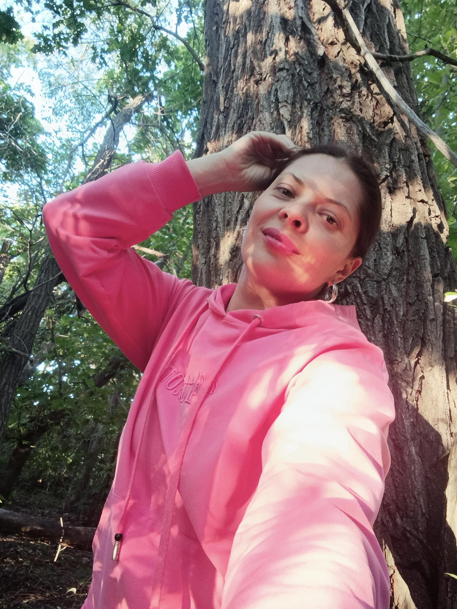 A low-angle, eye-level selfie shows a woman in a bright pink hoodie leaning against a large tree trunk in a wooded area. The woman has short, dark hair, dark eyes, and is wearing pink lipstick. She is looking at the camera with her right arm raised and her hand resting on her head. She is also wearing large, hoop earrings. The tree trunk is rough and textured, with sunlight dappling its surface. The background consists of green leaves and branches, suggesting an outdoor, natural setting. There is no visible indication of the city of Saratov, Russia.