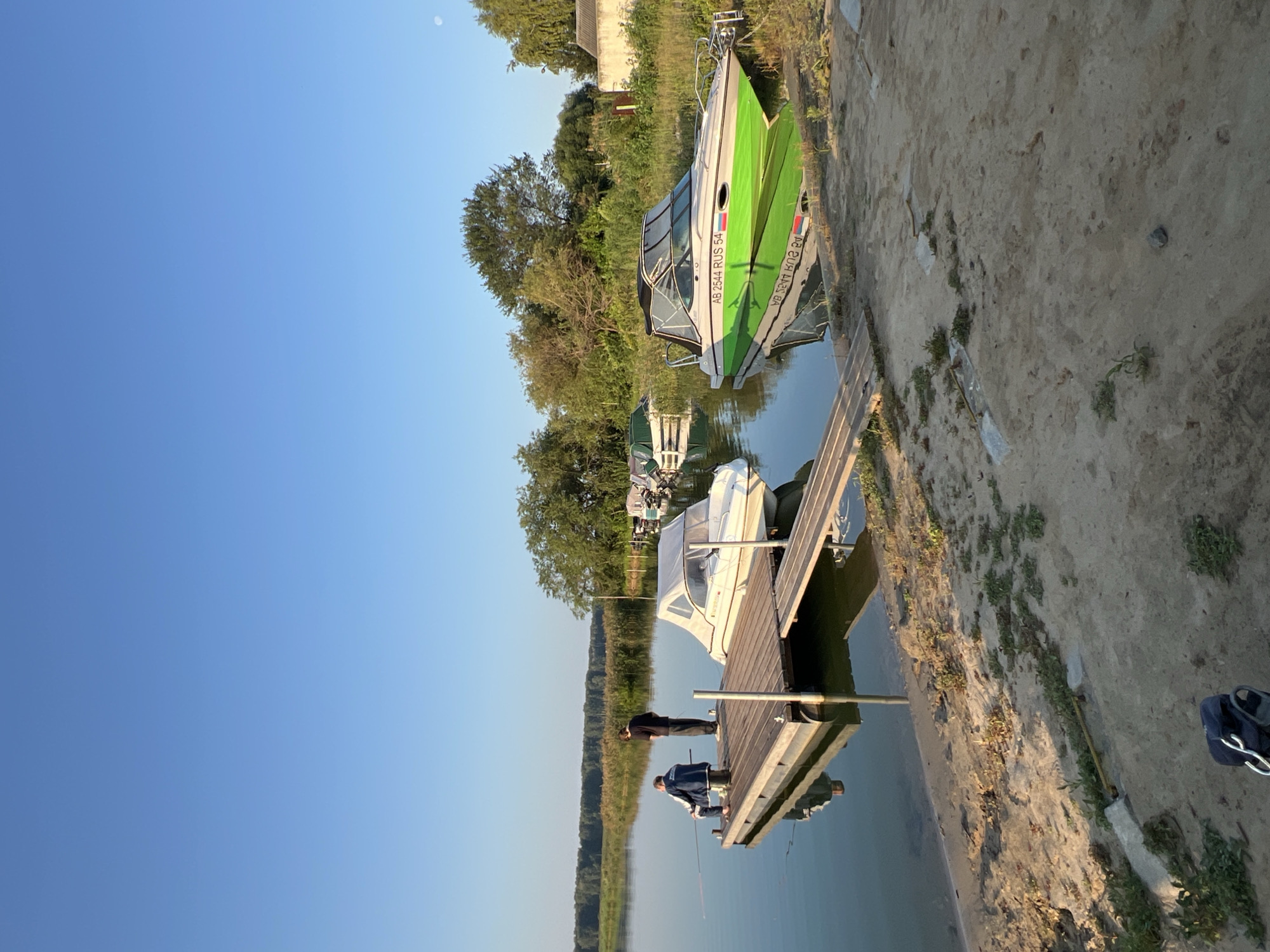 The image depicts a waterfront scene at a lake, likely in or near Novopichugovo, Russia, under a clear blue sky. The foreground shows a sandy bank with sparse vegetation and a dark bag lying on the ground. A wooden pier extends into the calm water, where two individuals are present. One person, seated and wearing a blue jacket and jeans, is fishing with a rod. Another person stands beside them. A white motorboat is docked at the pier. Further out on the water, several other boats are moored. One prominent boat in the mid-ground is lime green with a black canopy and displays the registration "AB 2544 RUS 54". The background features a dense line of trees and reeds bordering the lake, with some buildings visible on the right side of the frame. The overall impression is a tranquil, possibly recreational, setting during daylight hours.