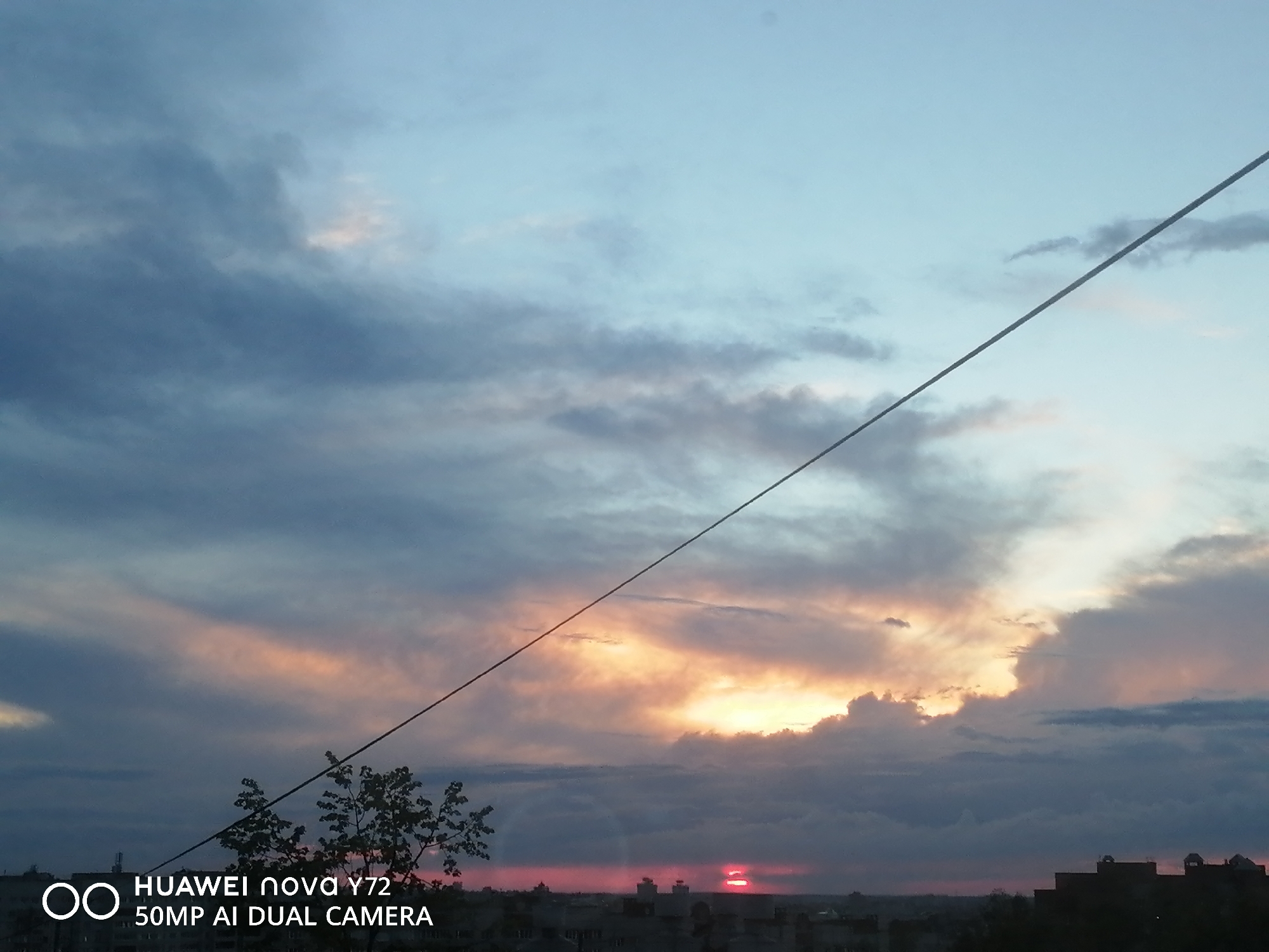 The image captures a vibrant twilight sky, likely a sunset, over what appears to be an urban or suburban landscape. The sky is filled with clouds, displaying a beautiful gradient of colors. The upper portion of the sky is a light, clear blue, transitioning downwards into a complex mix of dark blue, gray, and purple clouds. Near the horizon, these clouds are illuminated by the setting sun, revealing striking patches of bright orange, deep pink, and vivid red.
A prominent dark diagonal line, possibly a power line or cable, stretches across the sky from the upper left corner down towards the lower right. Below the horizon, the silhouettes of buildings forming a city skyline are visible, rendered in dark tones against the lighter sky. A few dark tree branches are also discernible in the lower left quadrant, further silhouetted against the colorful sky.
In the bottom left corner of the image, there is a watermark with white text: "OO HUAWEI nova Y72 50MP AI DUAL CAMERA", indicating the device used to capture the photograph. The overall mood is serene and dramatic, showcasing the natural beauty of the sky during twilight hours.