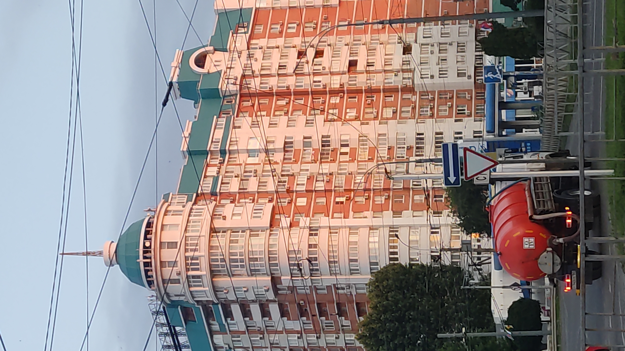 The image captures a street scene in Krasnodar, Russia, with a towering, modern residential building as the focal point. The building is a multi-story structure with a distinctive architectural style, featuring a curved facade with numerous windows and balconies. The facade is predominantly white with prominent horizontal bands of reddish-orange, giving it a striking appearance. A large, green, dome-like structure with a spire is visible at one end of the building, adding to its unique silhouette. Power lines crisscross the frame, suggesting an urban environment.
In the foreground, a red tanker truck is parked on the side of the road. The truck has a large cylindrical tank and appears to be a utility vehicle. Traffic signs are visible near the truck, including a yield sign and a directional sign. A blue and white structure that resembles a gas station or service point can be seen behind the truck and to the left. Trees and foliage are interspersed throughout the scene, indicating a well-maintained urban area.
The lighting suggests it is either early morning or late afternoon, with soft, warm light illuminating the building's facade. The sky is a pale, overcast blue. There is no clear indication of specific activity or event, but the presence of the truck and the signs imply a functioning street and infrastructure. The overall impression is one of a contemporary urban landscape in Russia. Some faint text is visible on the tanker truck, but it is not legible enough to discern. The license plate of the truck is also partially visible, with Cyrillic characters.