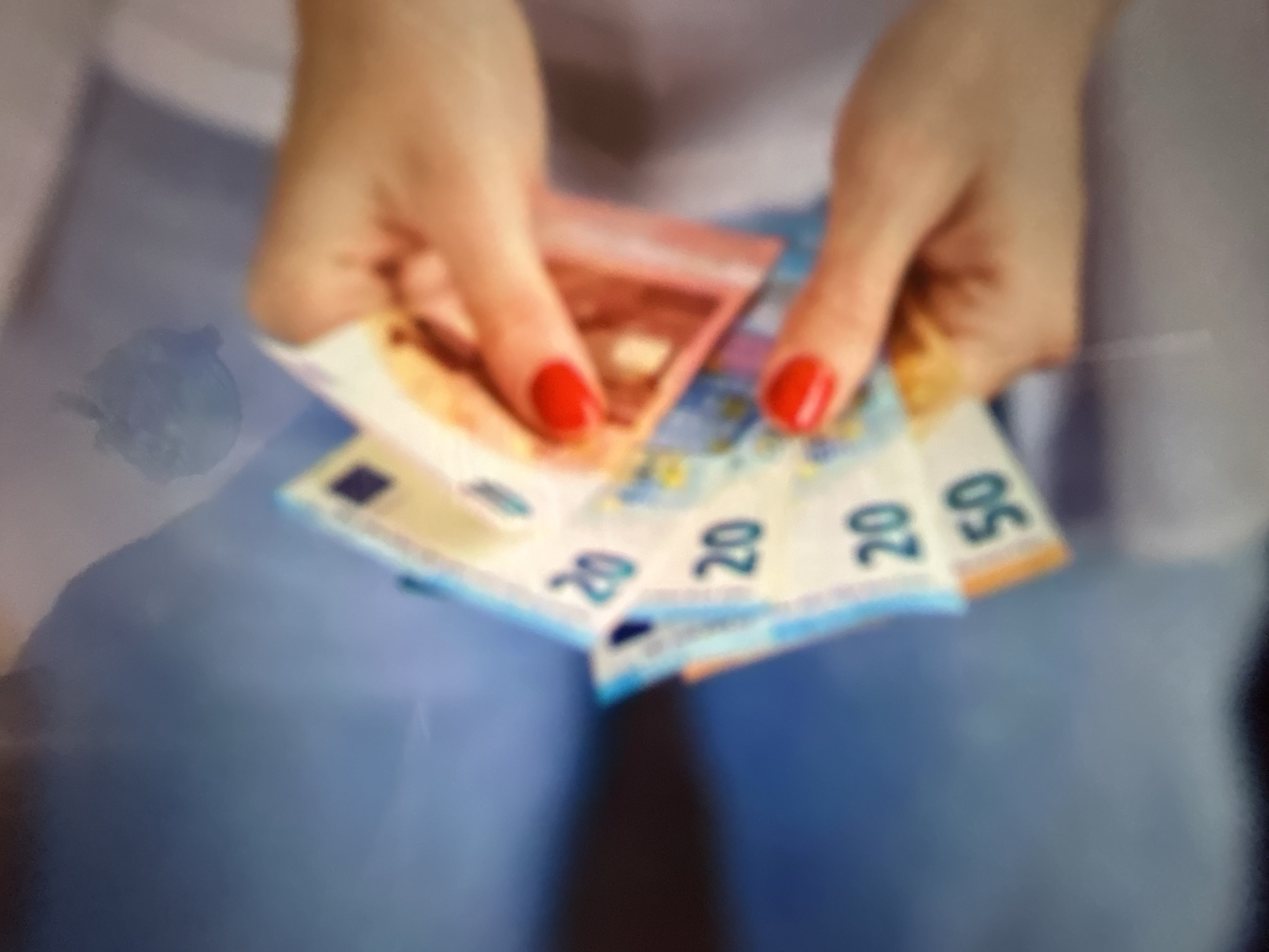 A person's hands, adorned with bright red nail polish, are holding several euro banknotes. The banknotes are fanned out and appear to be of denominations 20 and 50 euros, indicated by the visible numbers on them. The background is blurred and suggests an indoor setting with muted colors. The image lacks any specific indicators of location within Gelendzhik, Russia, or any discernible environmental details of the city.