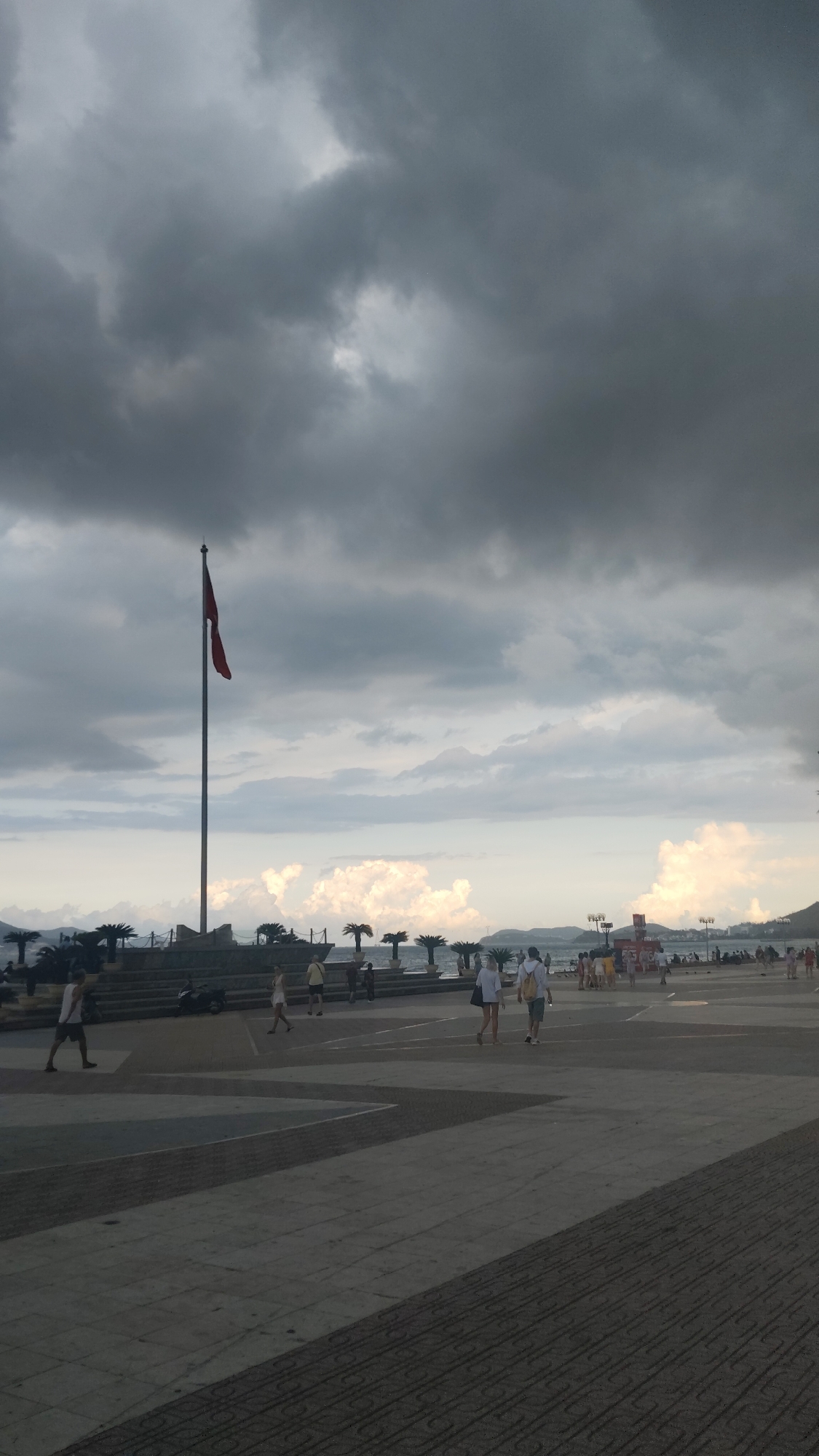 The photograph depicts a public square in Nha Trang Ward, Vietnam, under a heavy, overcast sky. Dominating the middle ground is a tall flagpole flying a red flag. Palm trees line a low concrete structure behind the flagpole, suggesting a waterfront location. People are sparsely scattered across the paved square, some walking in pairs and others alone. The paving pattern is geometric with alternating lighter and darker gray sections. In the distance, the shoreline and hints of buildings and structures can be seen, partially obscured by atmospheric haze. The overall mood is subdued, influenced by the dark sky.