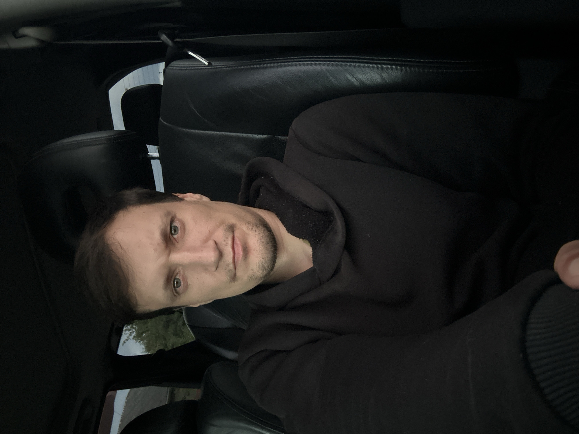 A man is seen from the chest up, seated in the passenger seat of a car. He has light skin, short brown hair, and green eyes. He is wearing a black hooded sweatshirt with a fuzzy grey lining visible around the hood. The interior of the car appears to be black leather. The background outside the car is blurry but suggests an outdoor environment with some greenery and structures. The overall lighting is dim, indicating it might be evening or a cloudy day. The image was captured in Makiv, Ukraine.