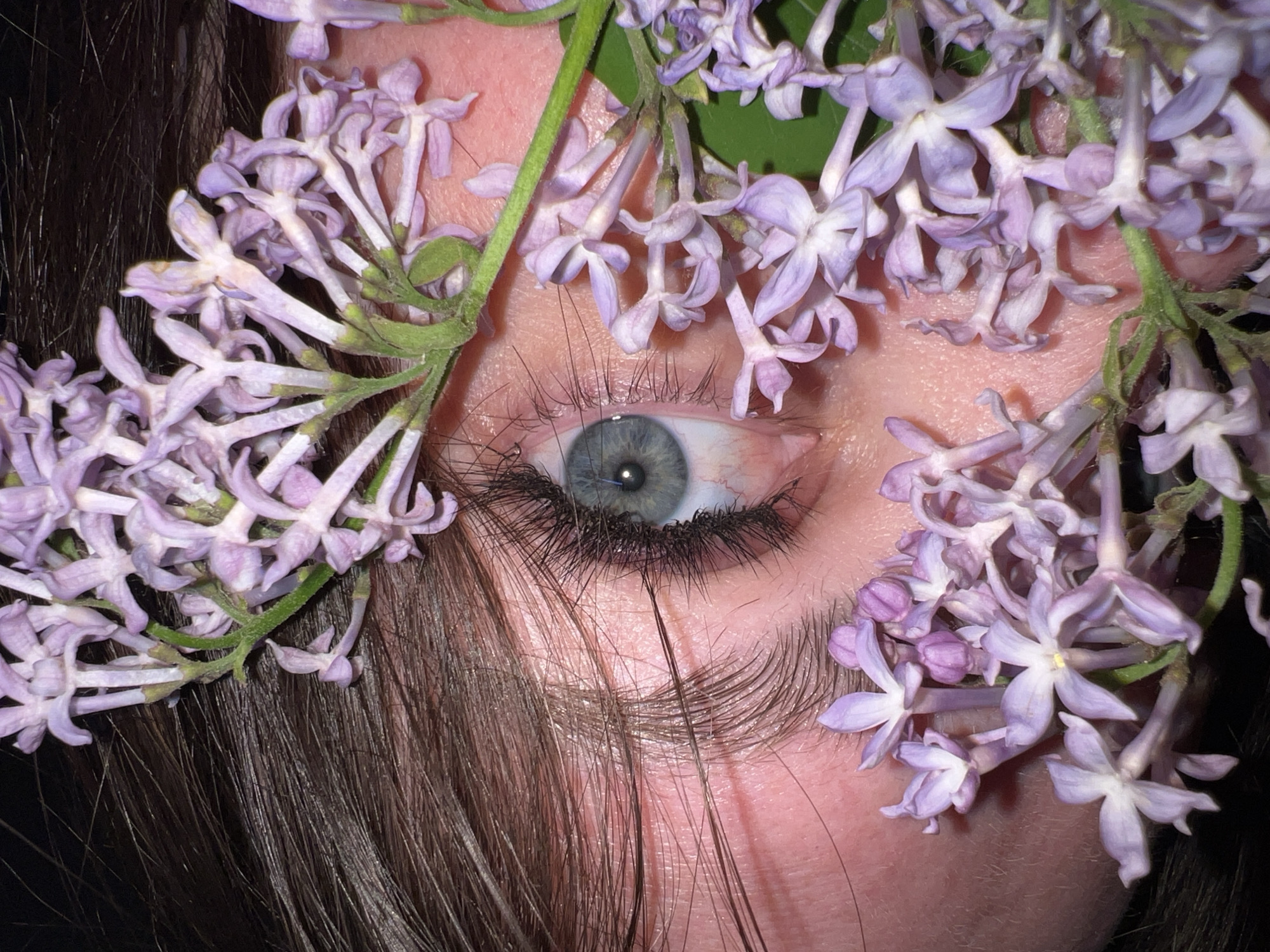 A close-up photograph focuses on a person's eye, surrounded by lilac flowers. The eye is light-colored with dark lashes. Strands of brown hair are visible near the eyebrow. The lilac flowers are light purple and arranged around the eye. The skin appears light and reflects the light. The photograph has a shallow depth of field, blurring the background. There is no evidence to determine the location of the city of Noviy, Russia.